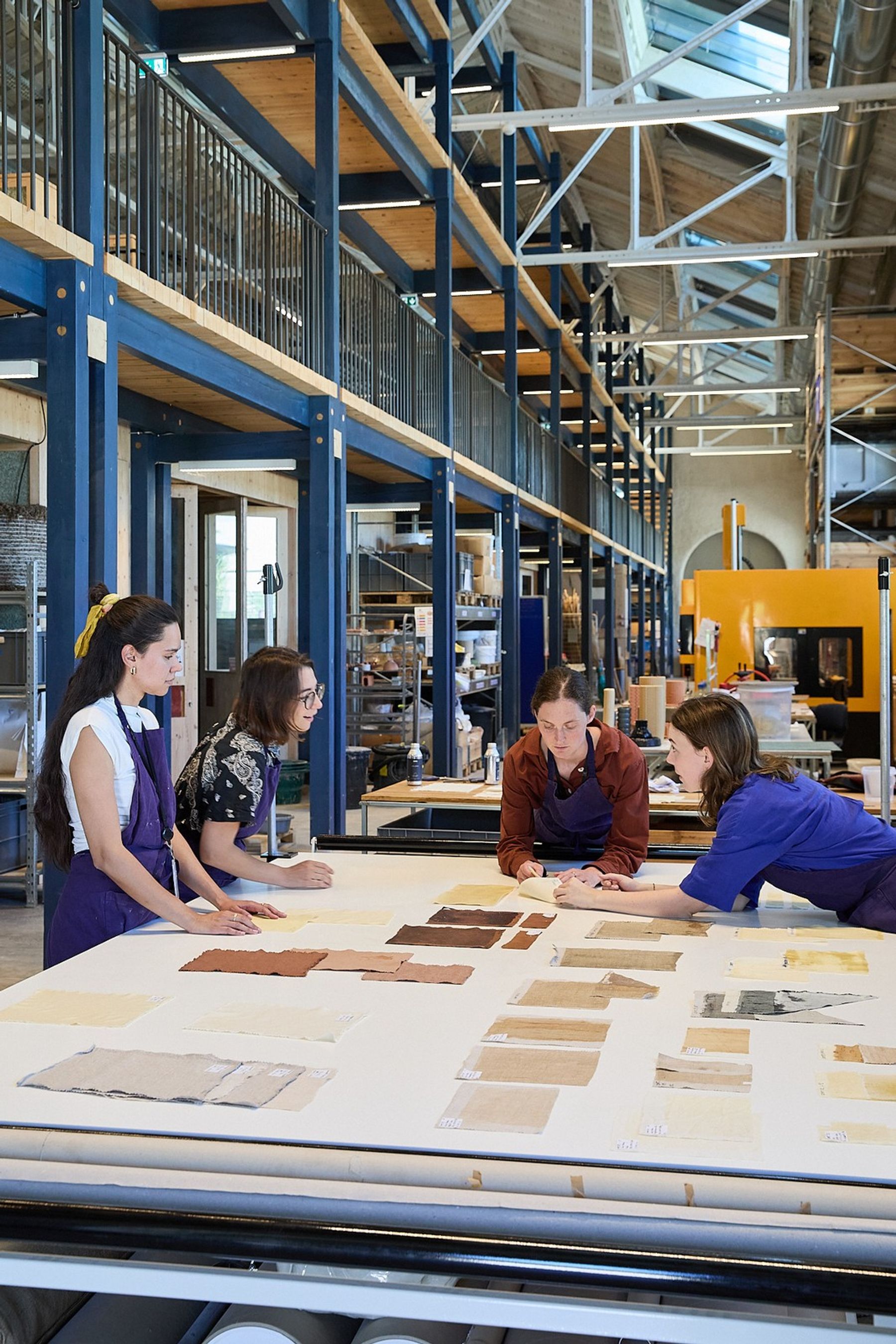 Four people in aprons gather around a large worktable in a spacious materials workshop, examining an array of textile and material samples in neutral and earth tones.