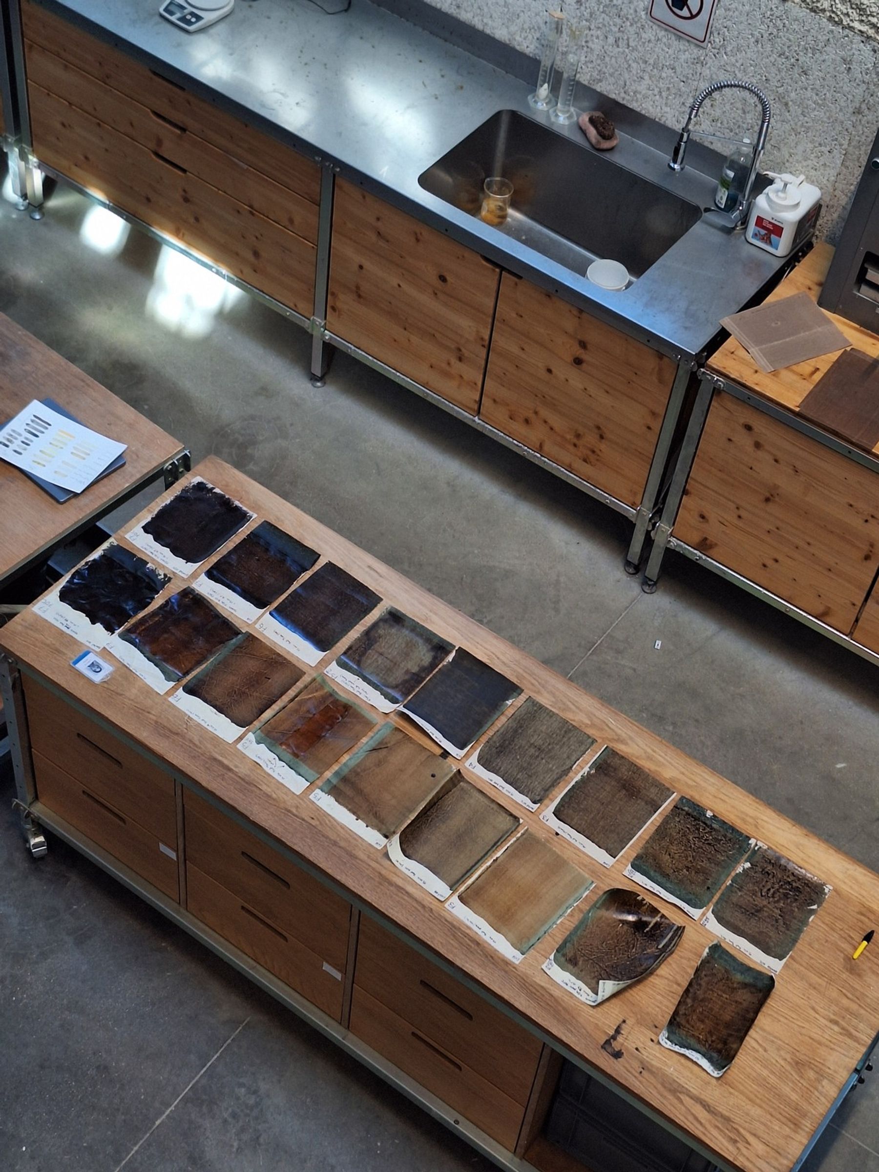 Overhead view of a materials lab with rows of dark brown, black, and earth-toned material samples arranged on a wooden worktable beside a stainless-steel sink and laboratory workstations.
