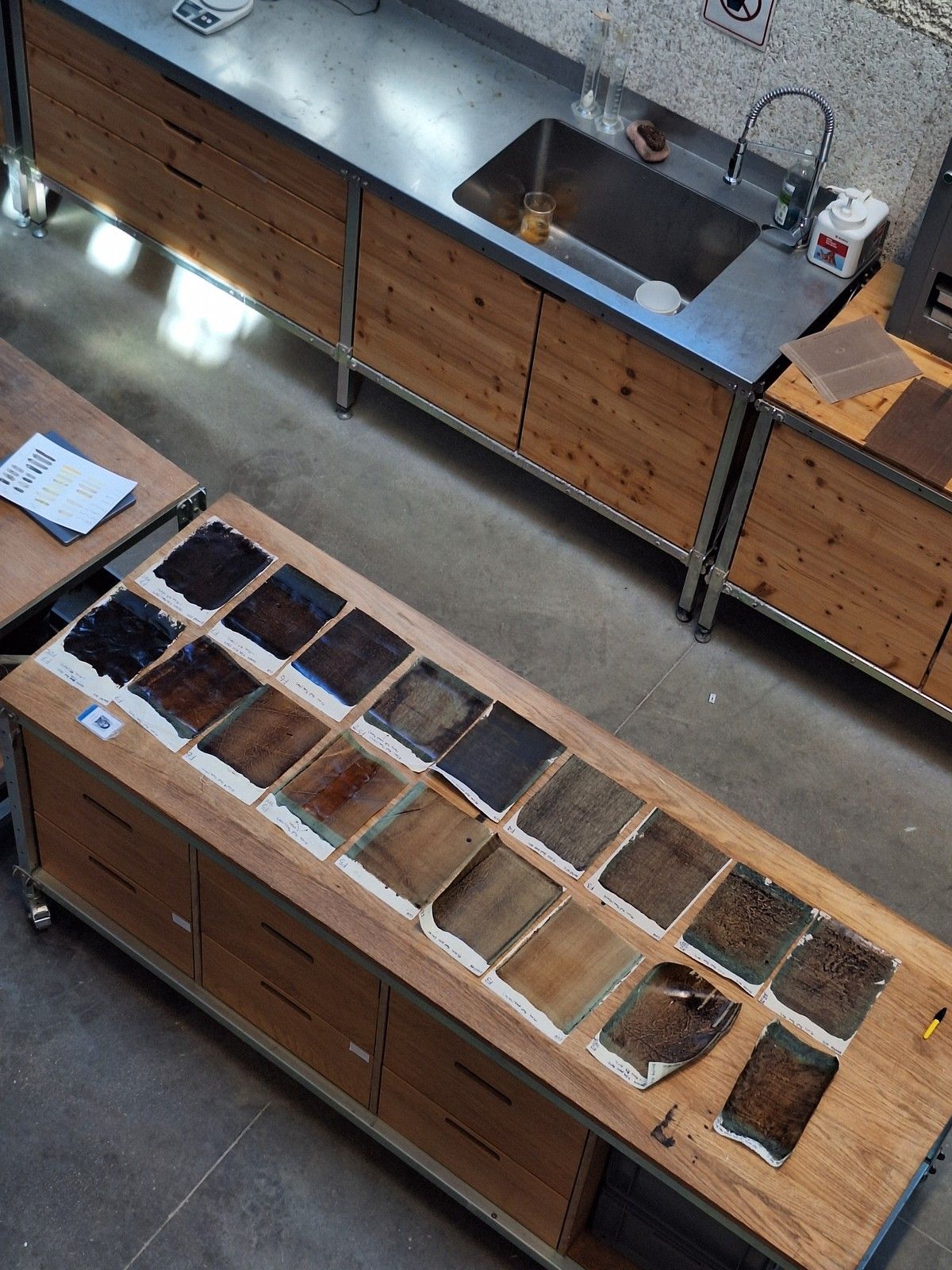 Overhead view of a materials lab with rows of dark brown, black, and earth-toned material samples arranged on a wooden worktable beside a stainless-steel sink and laboratory workstations.