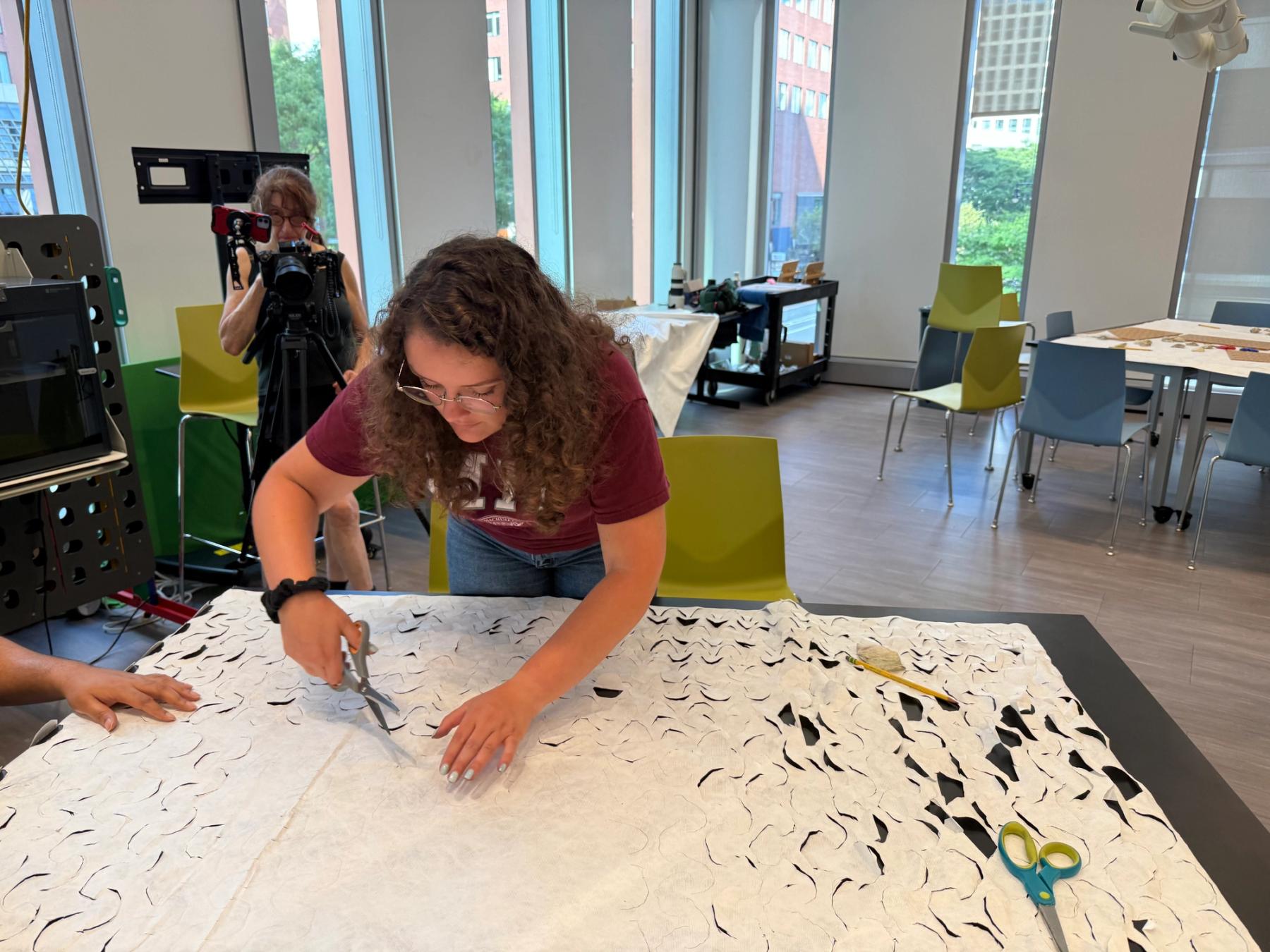 A person cuts repeating curved shapes into a large sheet of white textile while another person photographs the process in a design workshop.