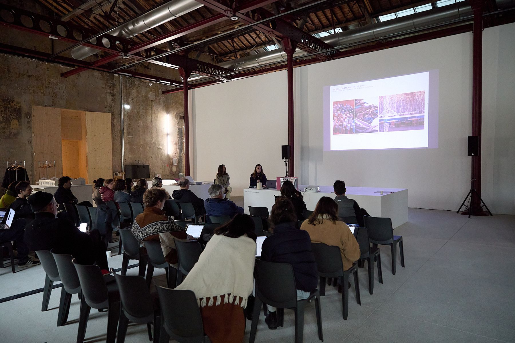 An audience attends a presentation in a large industrial space, with two speakers seated beneath a projected slide about natural colors and materials.