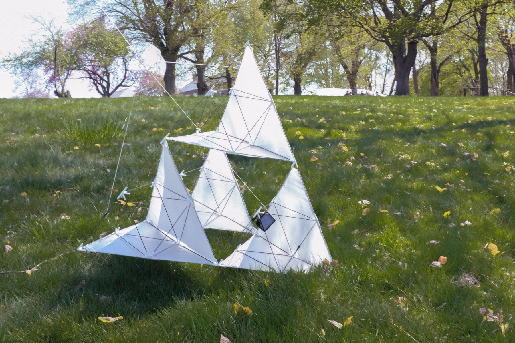 A lightweight geometric structure made from translucent fabric stretched over triangular frames in a grassy field.