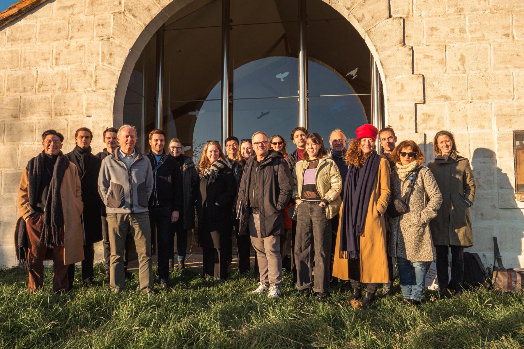 A group poses together outdoors in warm sunlight in front of a stone building with a large arched window.