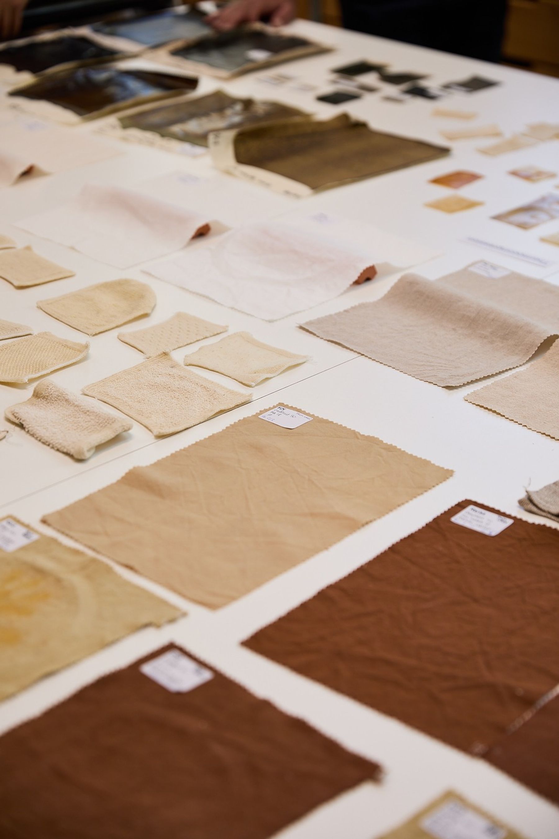 Close-up of assorted textile and material samples in cream, beige, brown, and black arranged across a white worktable, showing a range of textures, finishes, and thicknesses.