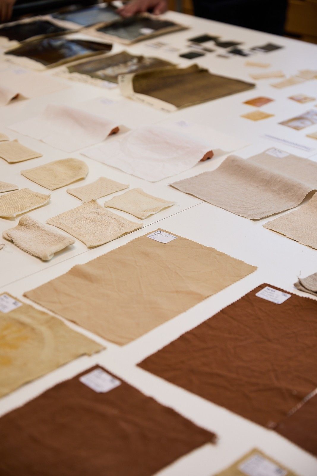 Close-up of assorted textile and material samples in cream, beige, brown, and black arranged across a white worktable, showing a range of textures, finishes, and thicknesses.