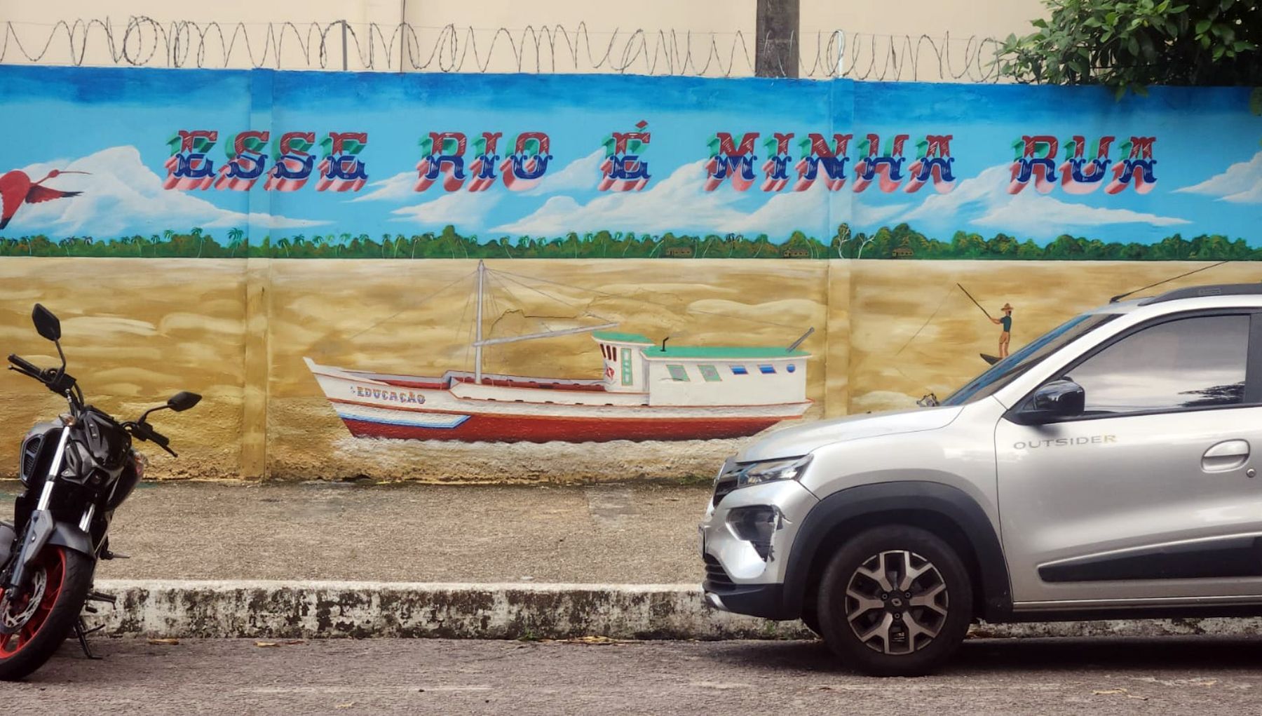A colorful mural depicts a river landscape with a boat and the Portuguese phrase “Esse rio é minha rua,” partially obscured by parked vehicles.