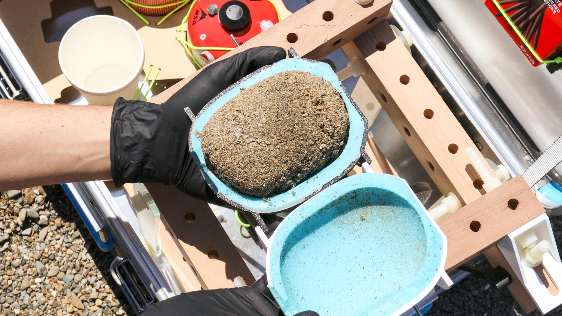 Gloved hands hold an open blue mold containing a compacted, sand-like material above a wooden fabrication apparatus.
