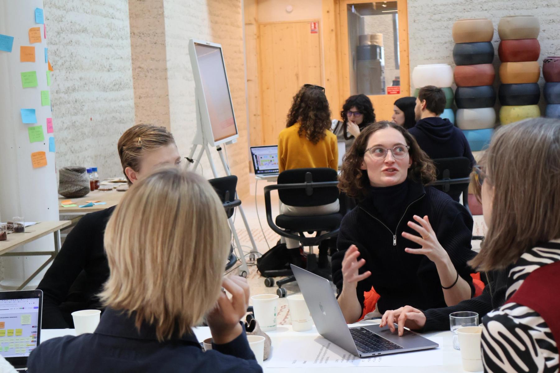 Workshop participants gather around a table with laptops, discussing ideas in small groups surrounded by digital displays and colorful notes.