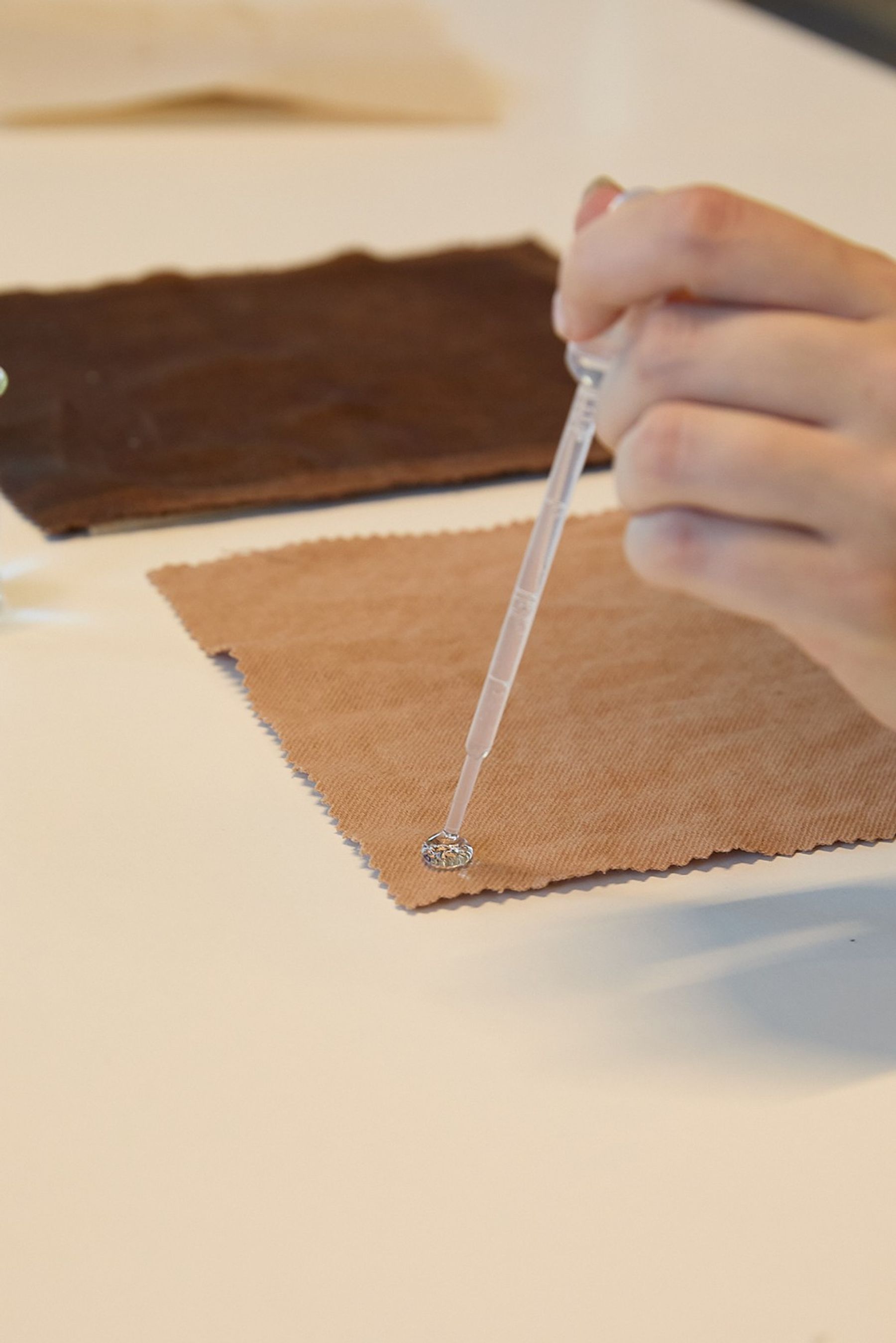 A hand uses a pipette to apply a clear liquid to a small sample on a square of brown fabric.