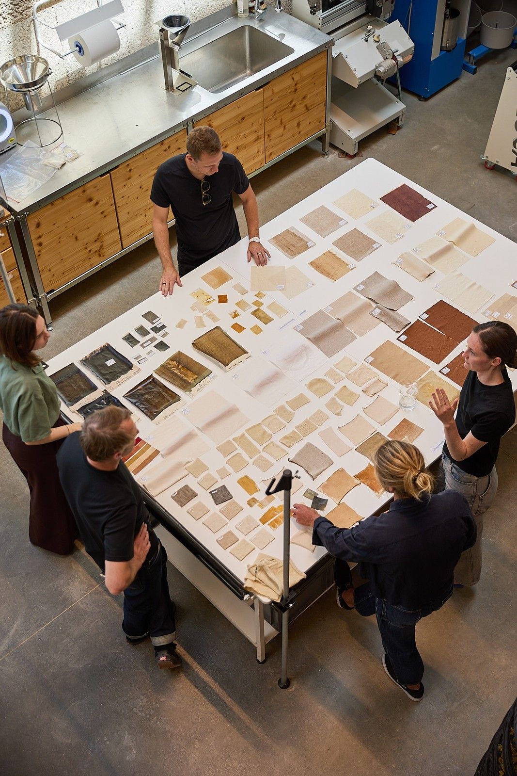 Five people gather around a large worktable in a materials lab, examining and discussing an array of textile and material samples in varied colors, textures, and sizes.