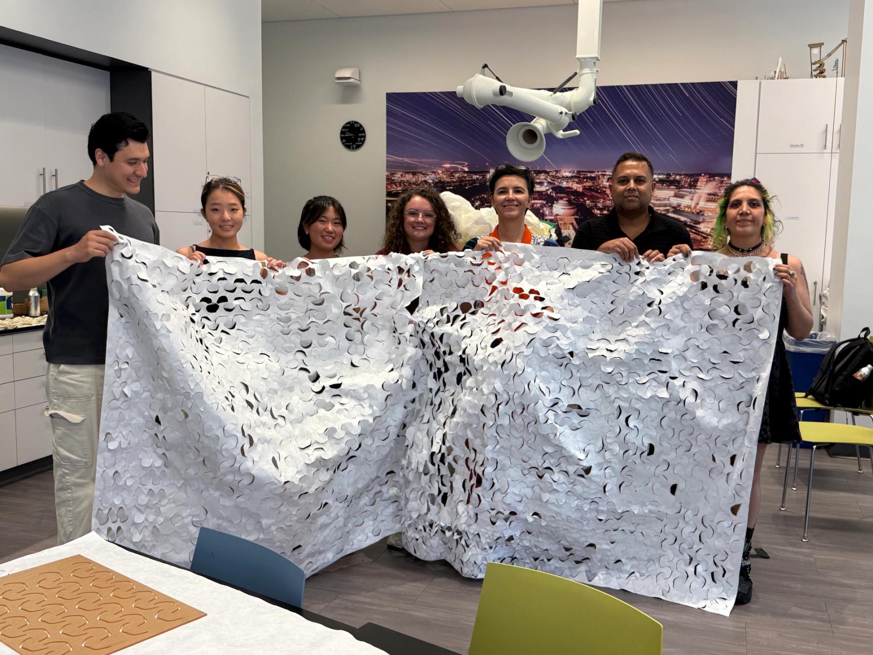 Seven people stand together in a design workshop holding a large white textile sheet covered with repeating curved cutouts.