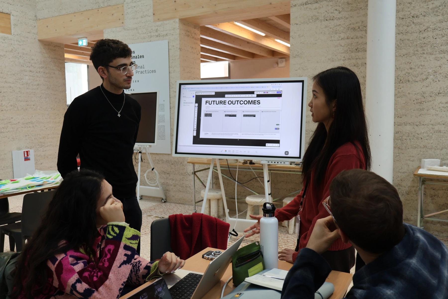 Workshop participants discuss ideas around a table, with one participant presenting beside a digital screen displaying “Future Outcomes.”