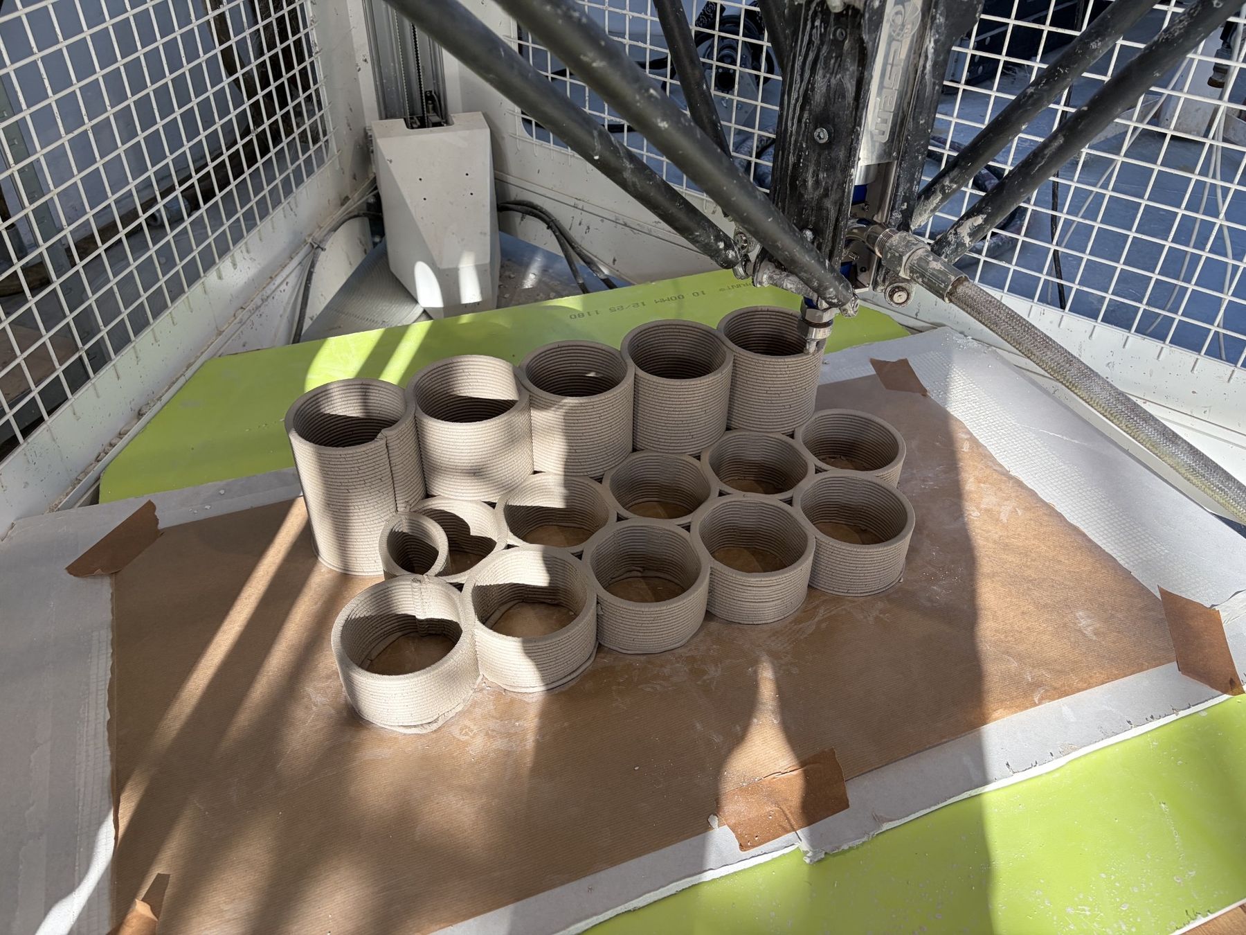 A robotic 3D printer fabricates a cluster of cylindrical, hollow structures in layered material on a flat work surface.
