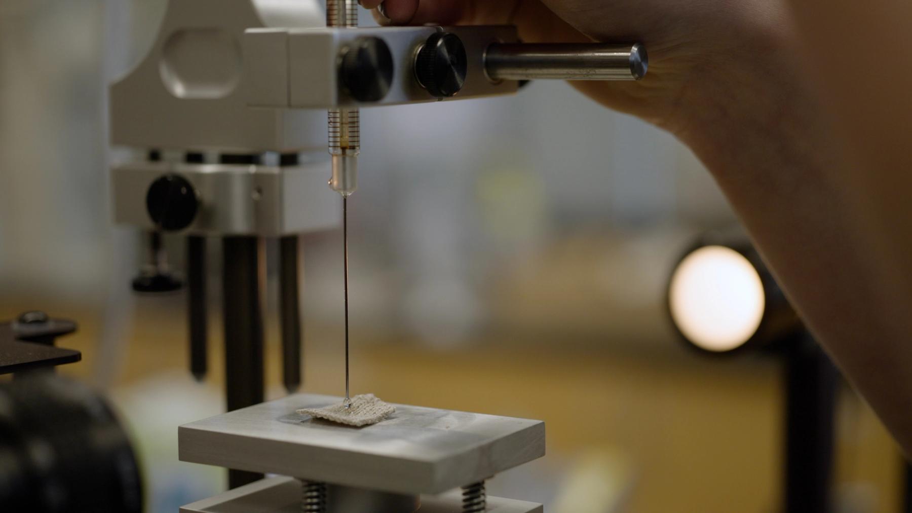 Close-up of a laboratory apparatus lowering a fine needle onto a small textured material sample positioned on a testing platform.