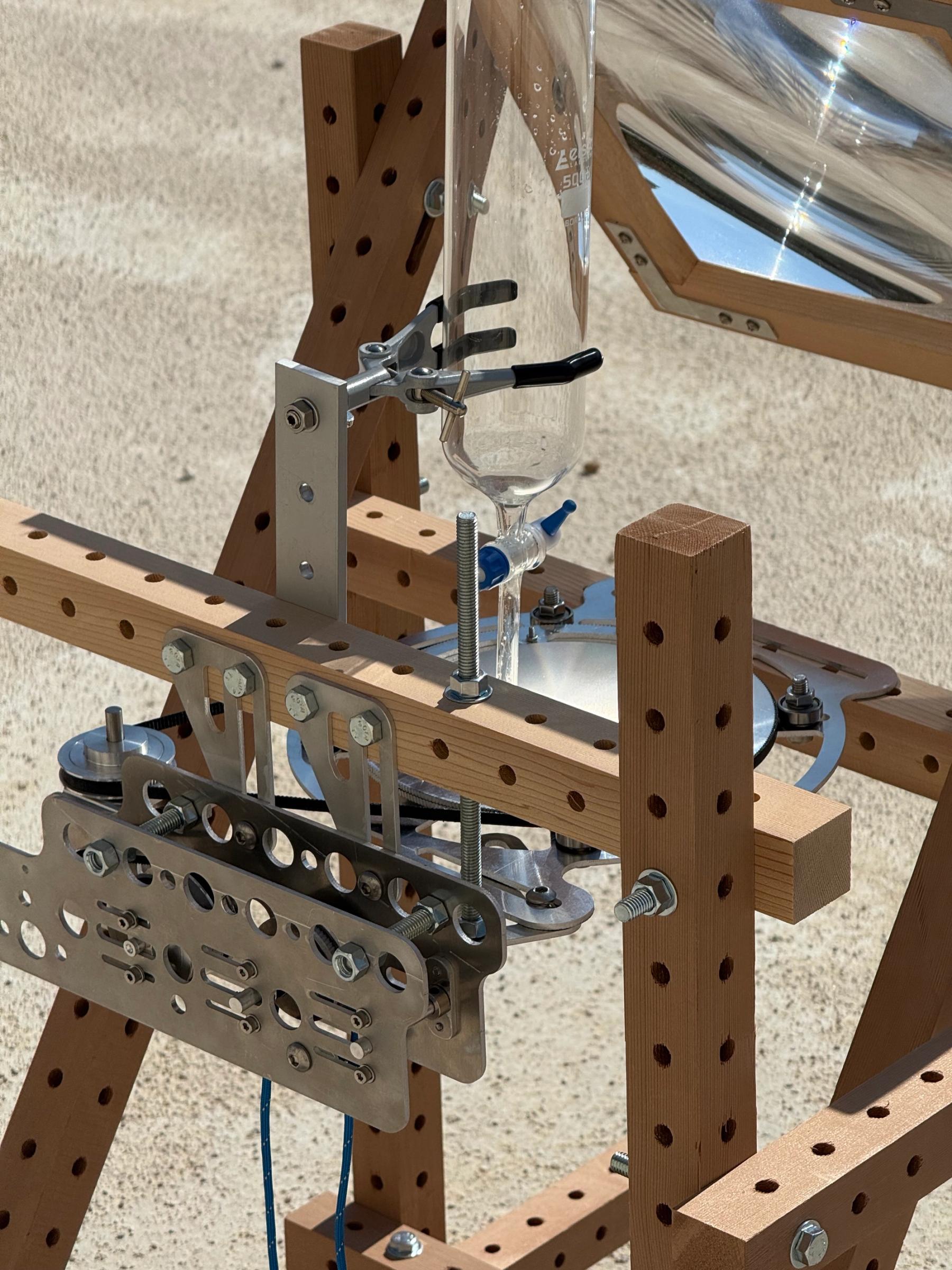 Close-up of an experimental apparatus made from a perforated wooden frame, metal components, glassware, and a reflective dish.