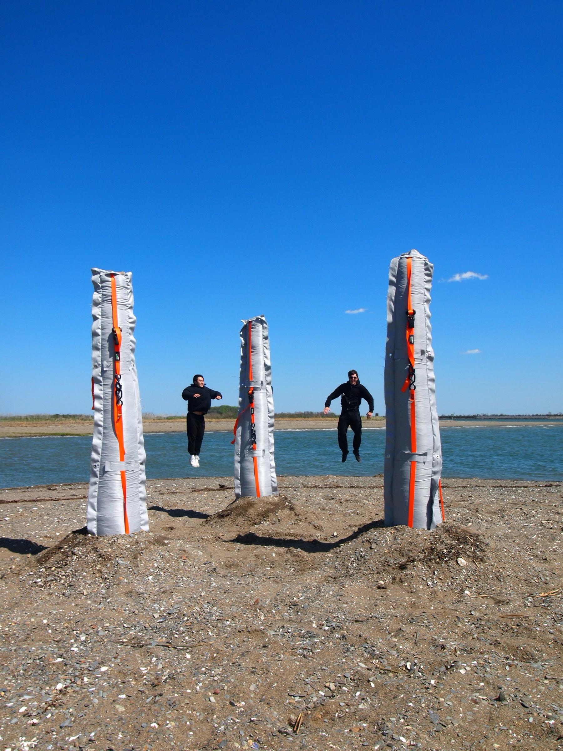 Two people jump between three tall, fabric-wrapped structures with bright orange straps installed on a sandy shoreline beside the water.