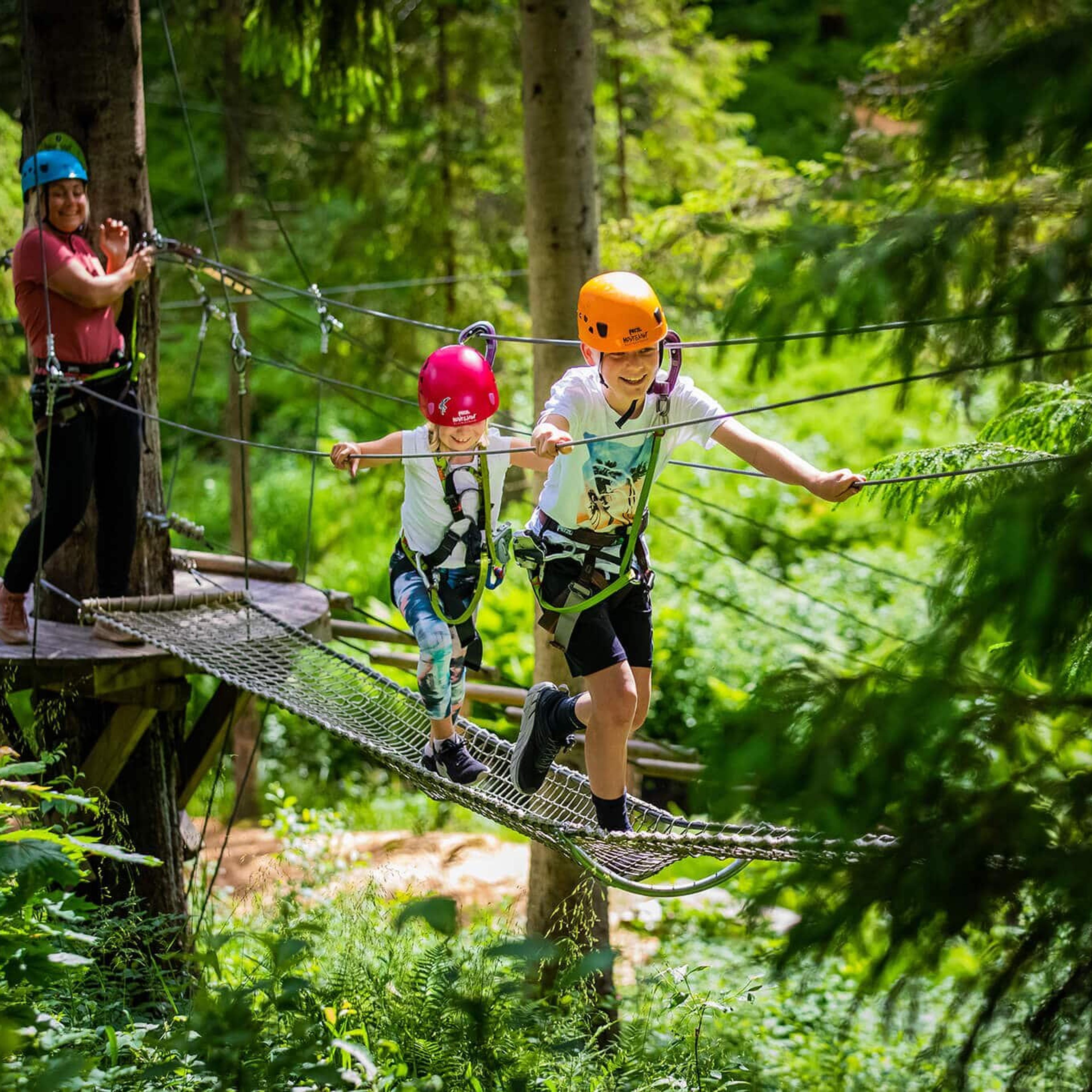 Barn med hjelm og klatresele balanserer på en hengebro i en klatrepark i skogen