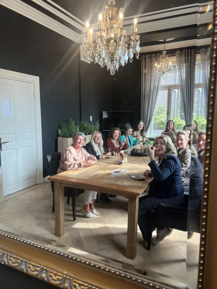 Groep vrouwen aan een lange tafel in een ruimte met kroonluchters, weerspiegeld in een gouden spiegel