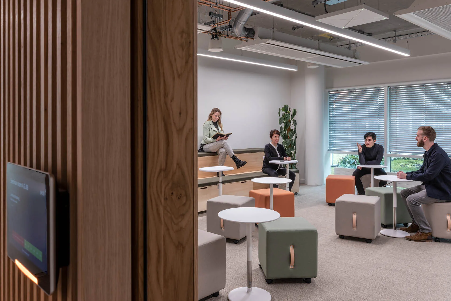 Staff sit on bleacher seating and square stools in a room with a digital booking display on the door