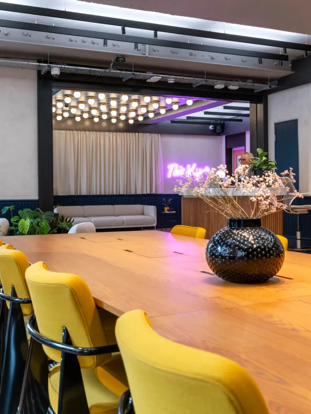 A vase of dried flowers on a long meeting table surrounded by yellow chairs