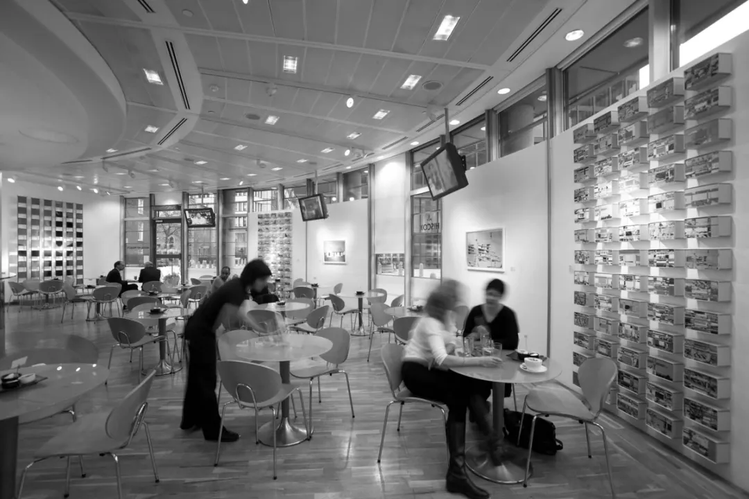 Black and white photo of the original office cafe with tables and chairs, and clusters of small artworks on the curved wall