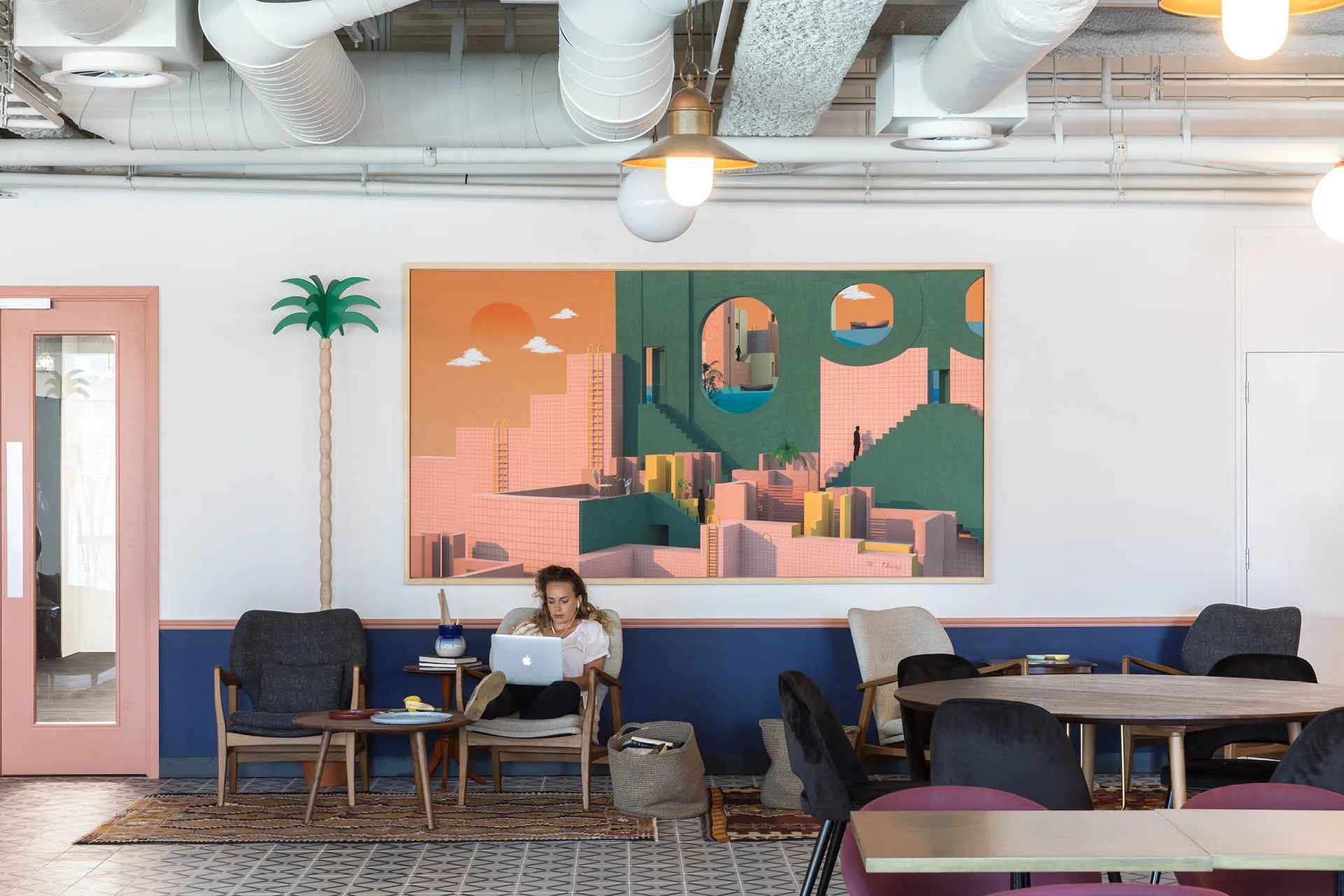 A woman works on a laptop from an armchair with a large colourful painting behind and casual tables to the side