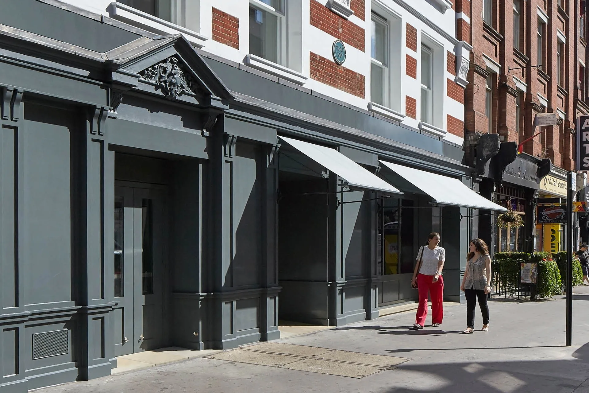 Black painted stone building entrance with 2 canopies over windows on to street. Two figures walking in front of building. 