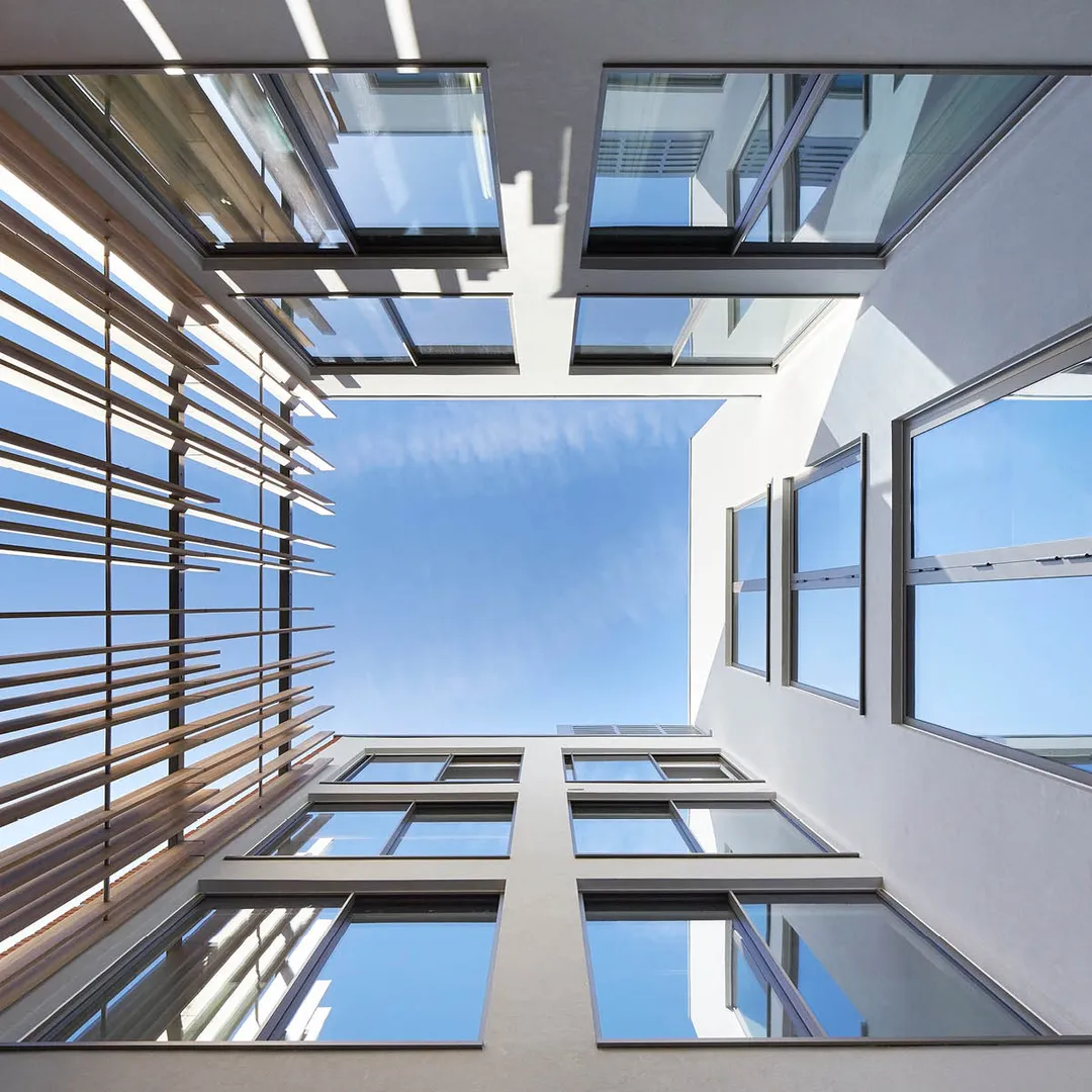 View of blue sky surrounded on 3 sides by flat white facades with large windows and irregularly spaces tall wooden louvres 