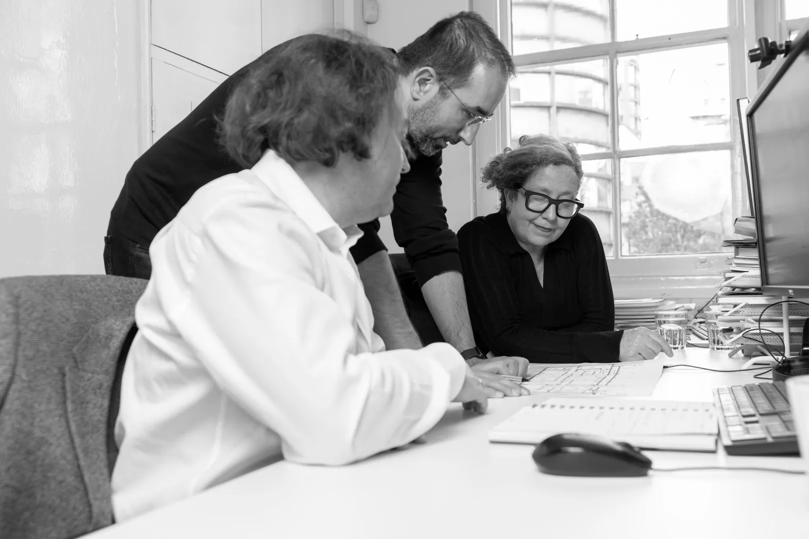 Black and white photograph of 3 people working together looking at plans on desk, with one person standing