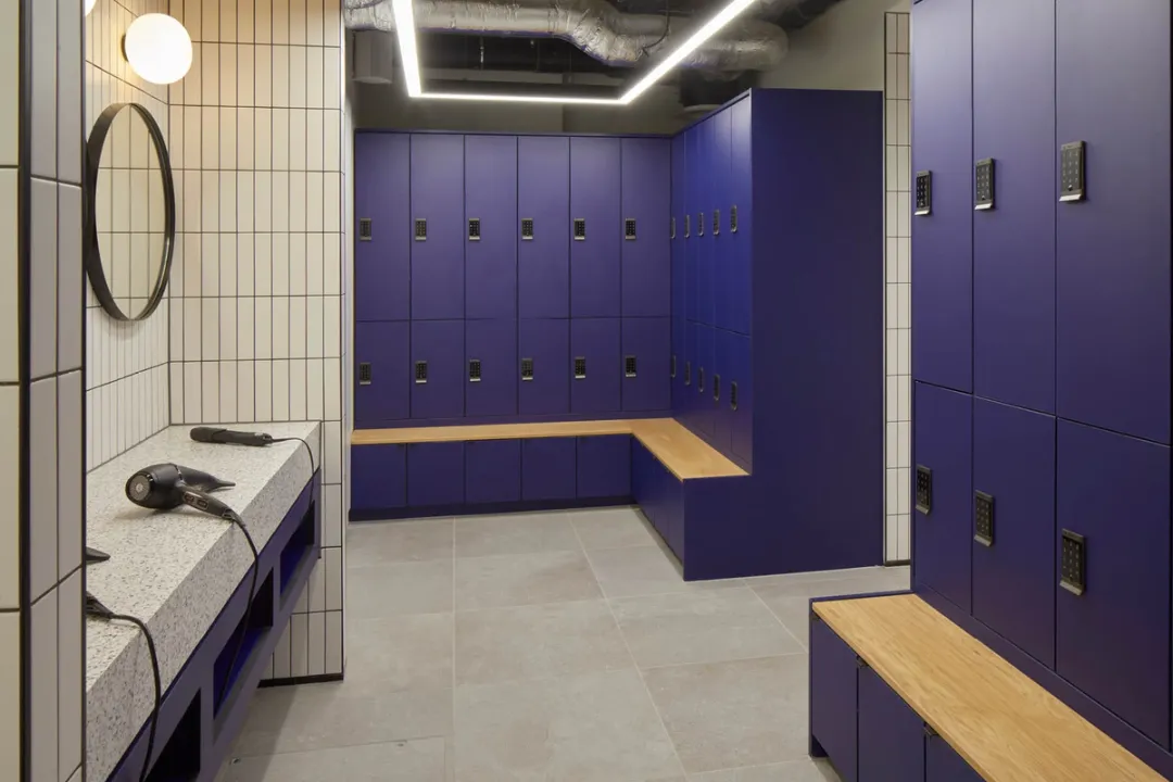 A changing room with a marble vanity with a mirror and hairdryers, tall navy blue lockers and wood bench seating