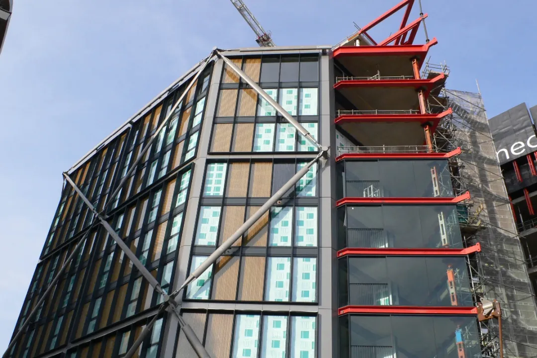 View upwards of glass tower construction with three upper levels awaiting glass walls, hoarding to right