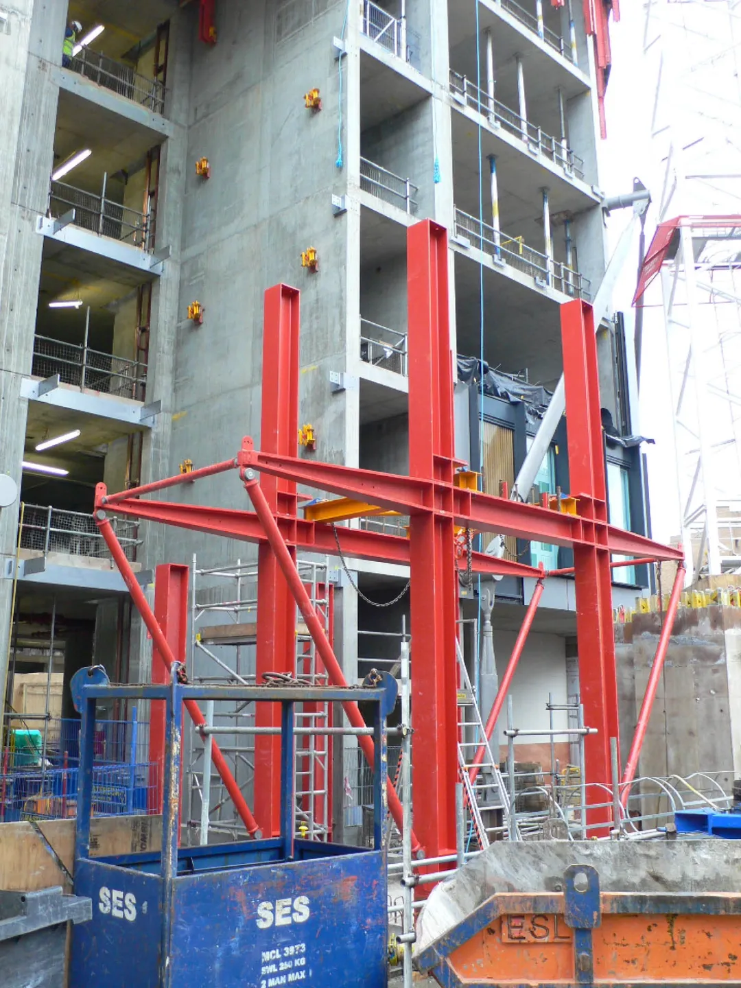 Close view of red steel girders and cross bracing half constructed next to concrete building shell