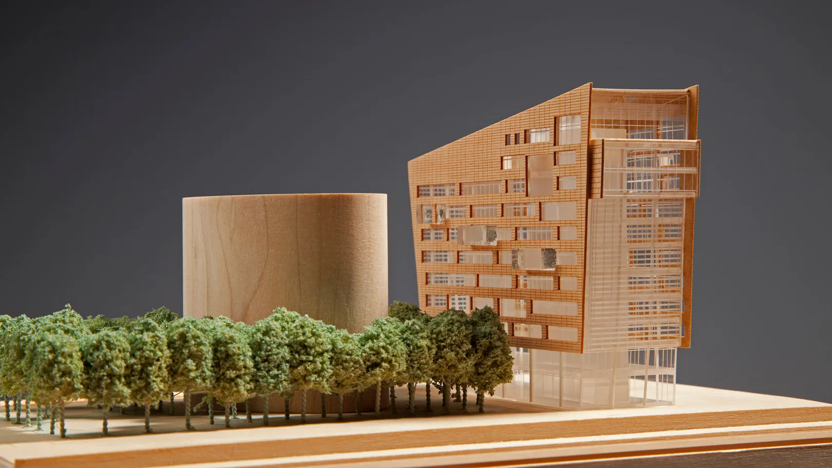 Wooden model of a building with trees in front of of modern building with angled roof with a cylindrical structure at rear and trees to the side