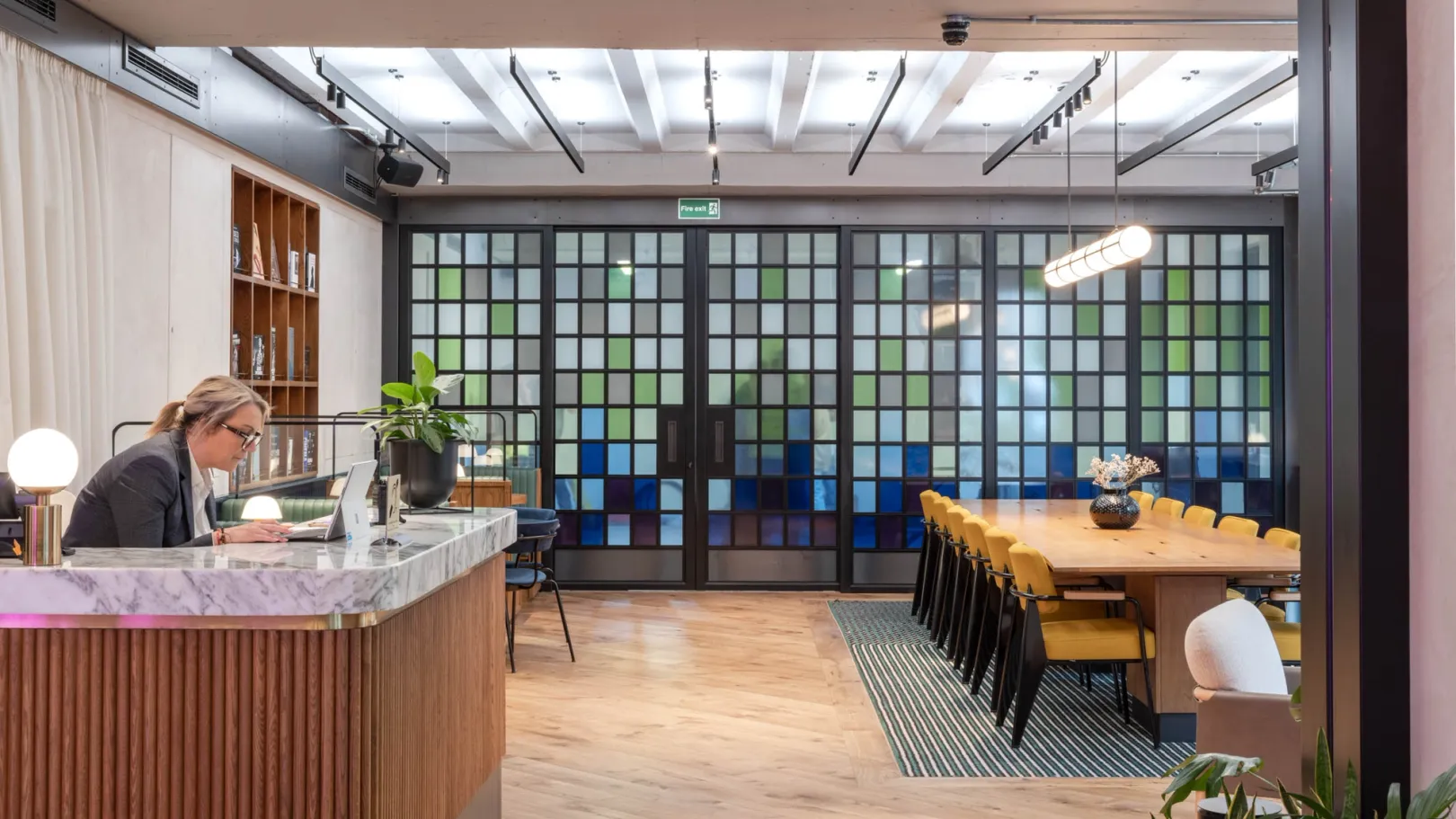 A receptionist sat at a wooden desk opposite a long meeting table, green and blue glass screen wall behind
