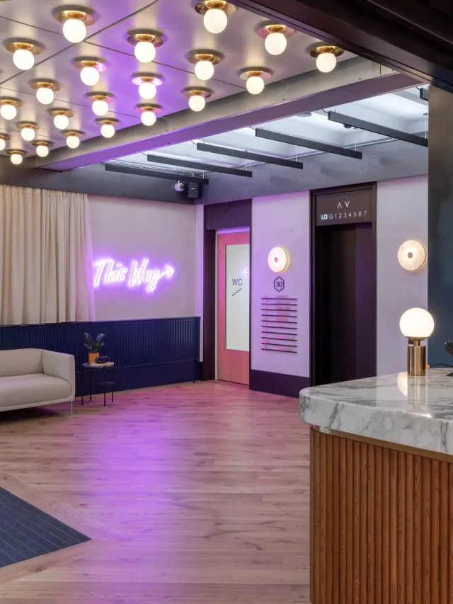 A neon 'this way' sign next to a door to stairs and a lift, round ceiling bulbs above