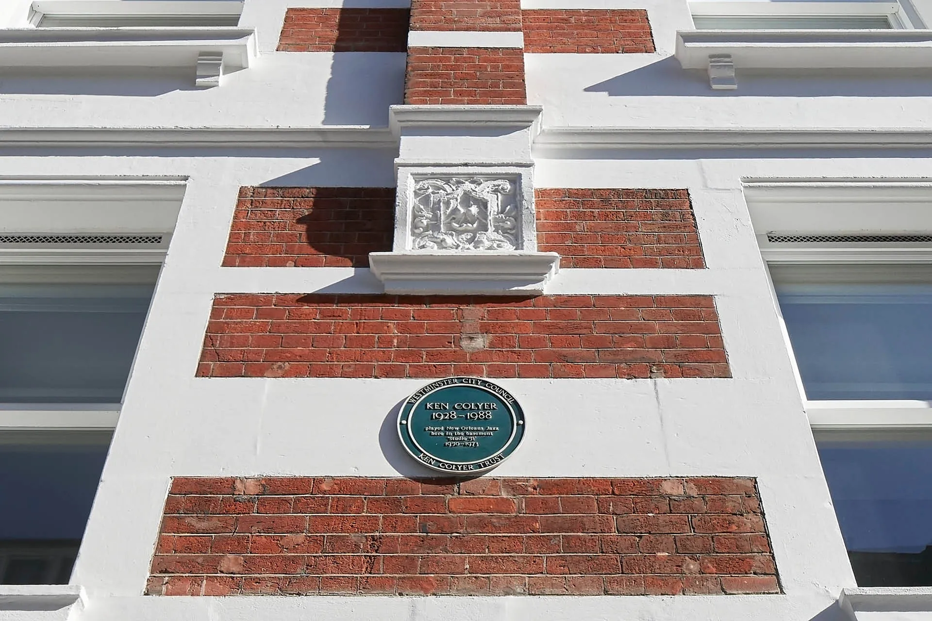 Close up of a brick facade with white painted window frames, a blue circular plaque at the centre reads 'Ken Colyer'
