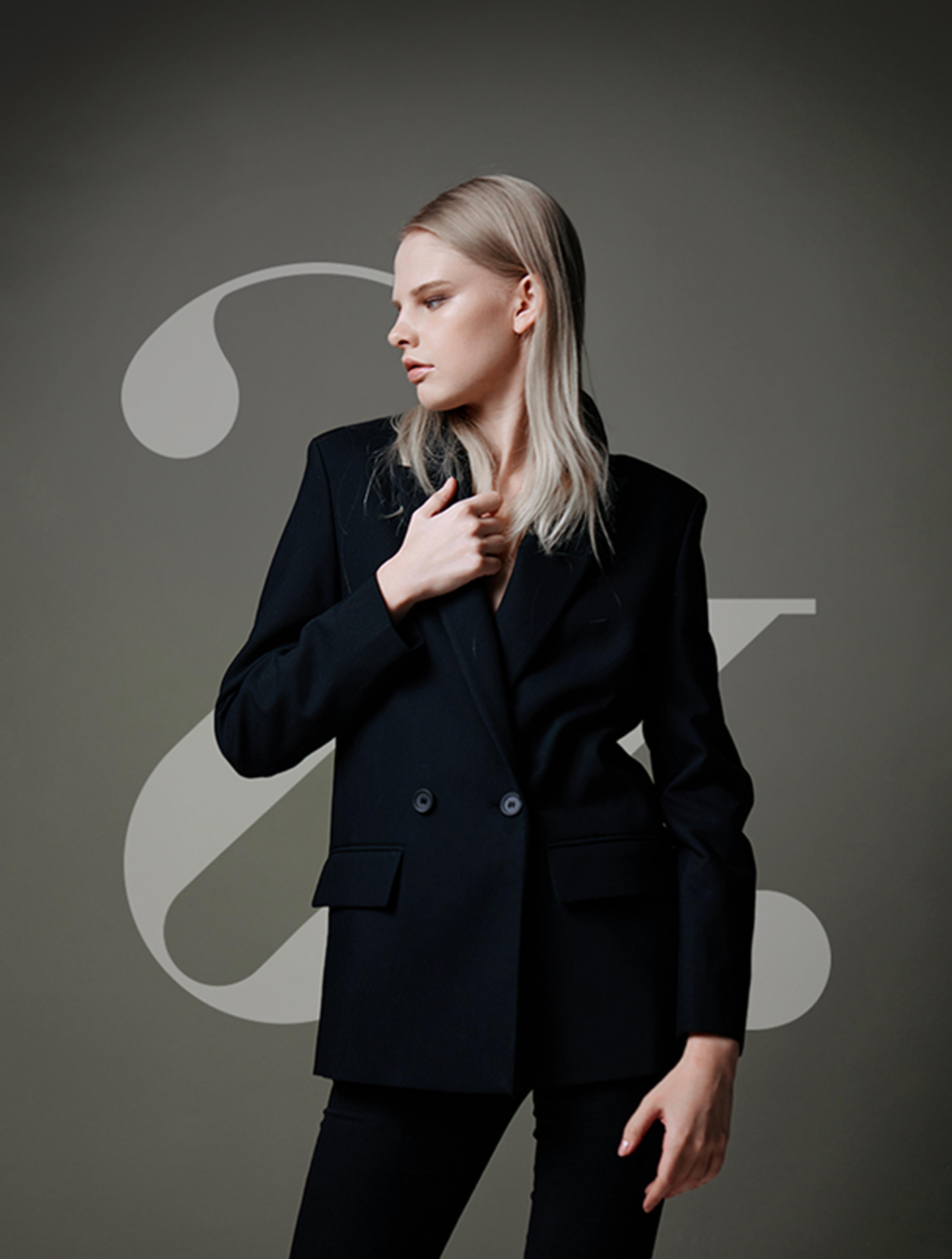 Eine elegante Frau in schwarzem Blazer posiert vor einem schlichten, dunklen Hintergrund mit geschwungenen Designelementen.