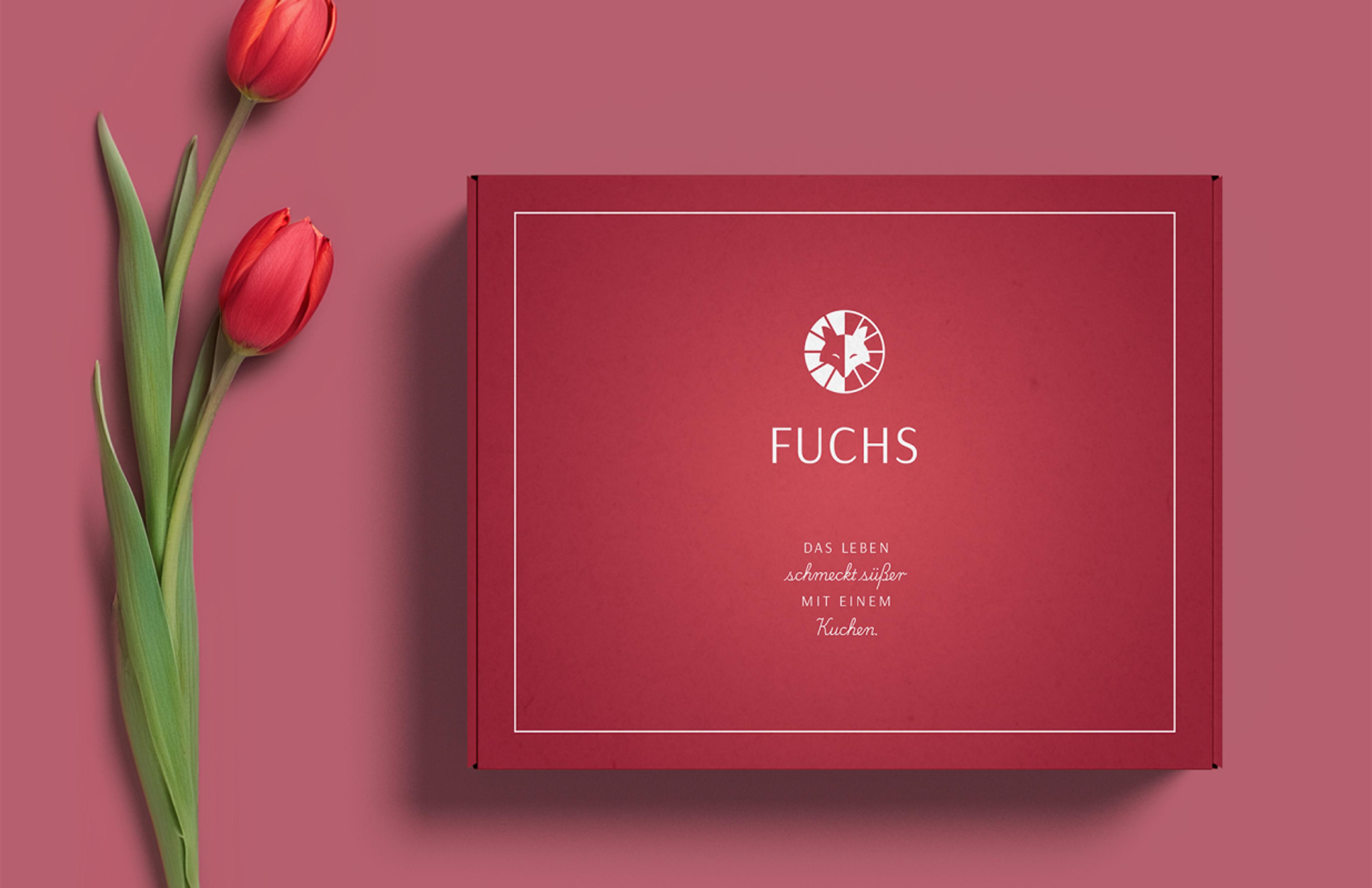Eine elegante, rot designte Verpackungsschachtel mit dem „Fuchs“-Logo und Slogan. Daneben liegen zwei rote Tulpen auf einem farblich abgestimmten Hintergrund.