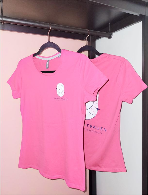 Zwei pinke T-Shirts mit dem Zauberfrauen-Logo, hängend an einer Garderobe. Das Design symbolisiert weibliche Stärke und den Kampf gegen Brustkrebs. - Unerwartet Design