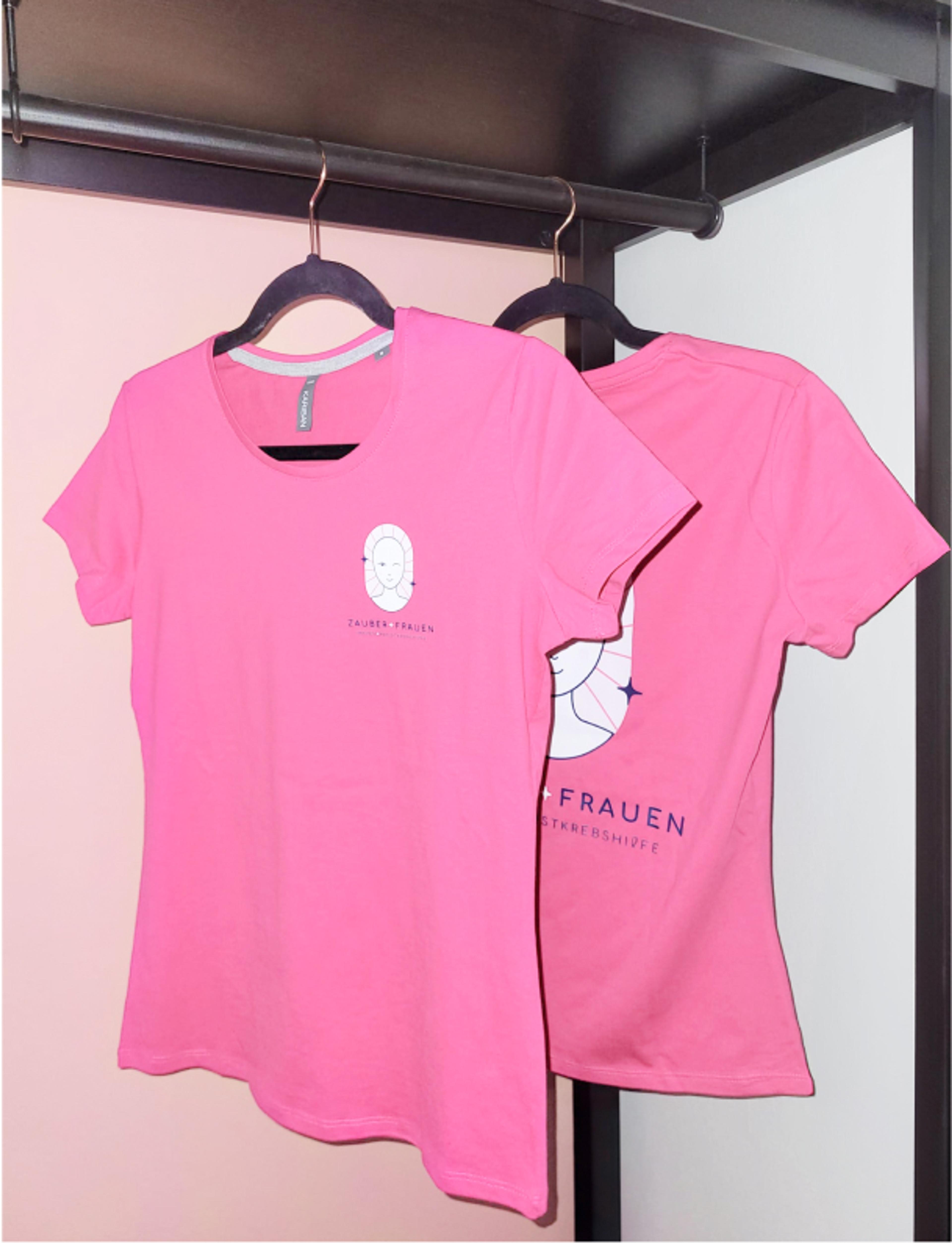 Zwei pinke T-Shirts mit dem Zauberfrauen-Logo, hängend an einer Garderobe. Das Design symbolisiert weibliche Stärke und den Kampf gegen Brustkrebs. - Unerwartet Design
