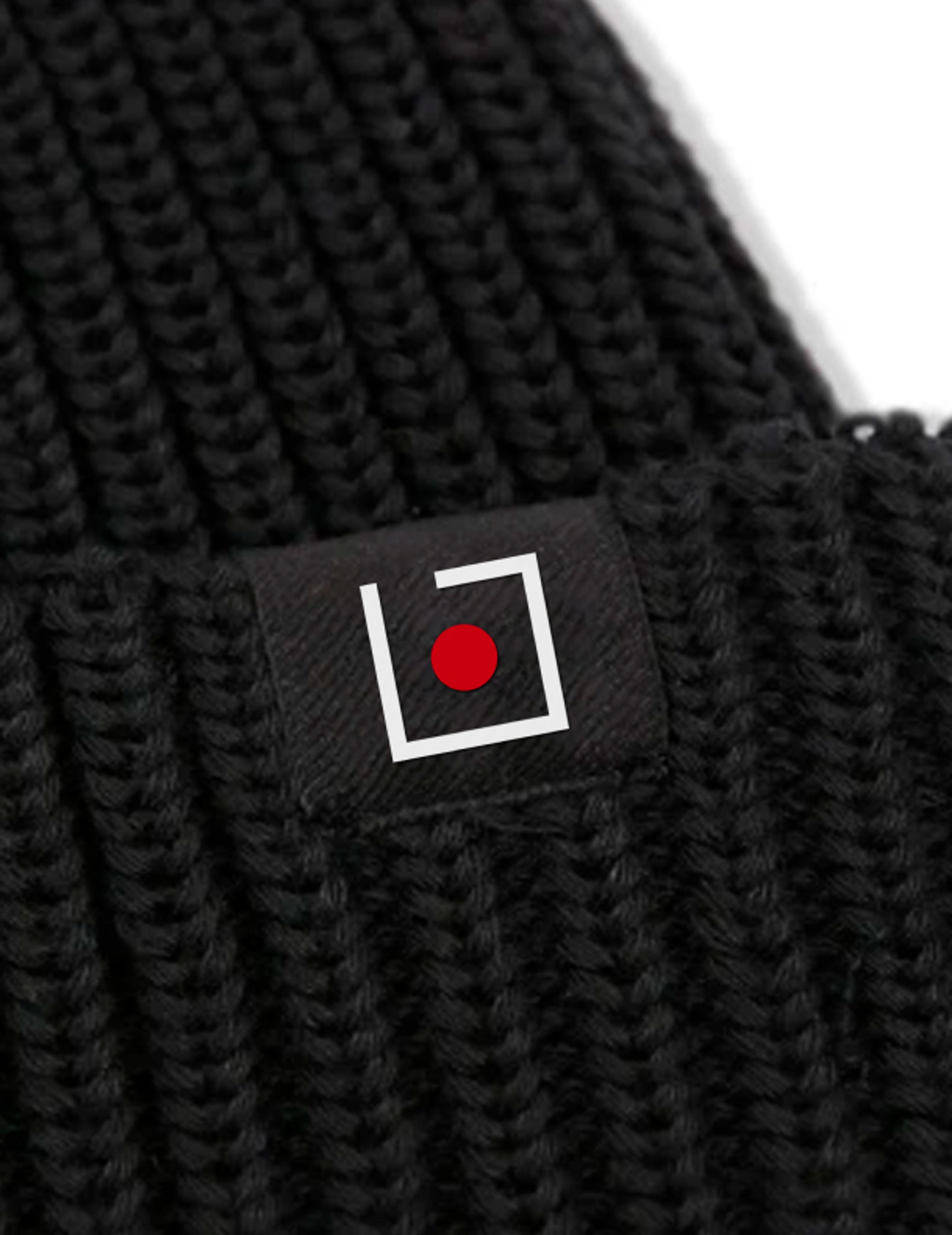 Ijob-Logo auf einer schwarzen Strickmütze – Eine Nahaufnahme eines schwarzen Beanies mit einem gestickten Ijob-Logo-Tag. Das Logo zeigt ein weißes Quadrat mit einem roten Punkt in der Mitte.