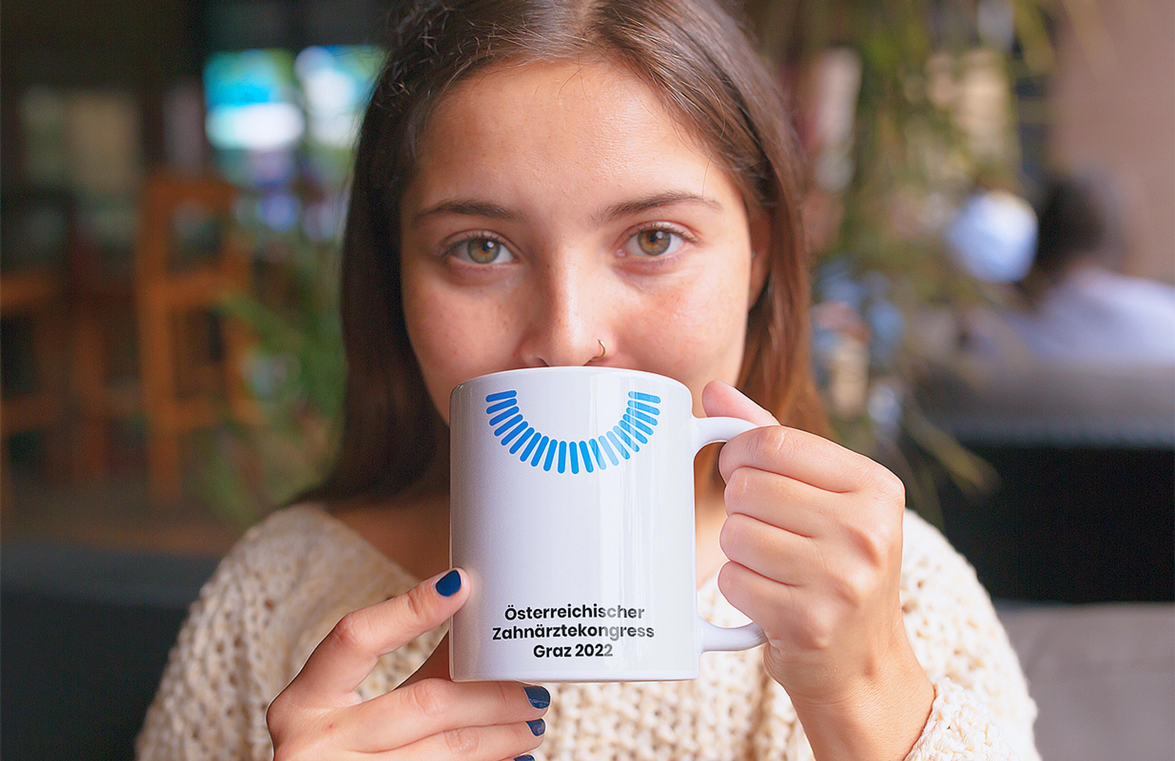 Junge Frau trinkt aus einer weißen Tasse mit dem blauen Halbkreis-Logo des Österreichischen Zahnärztekongresses Graz 2022, gestaltet von Unverwartet Design.