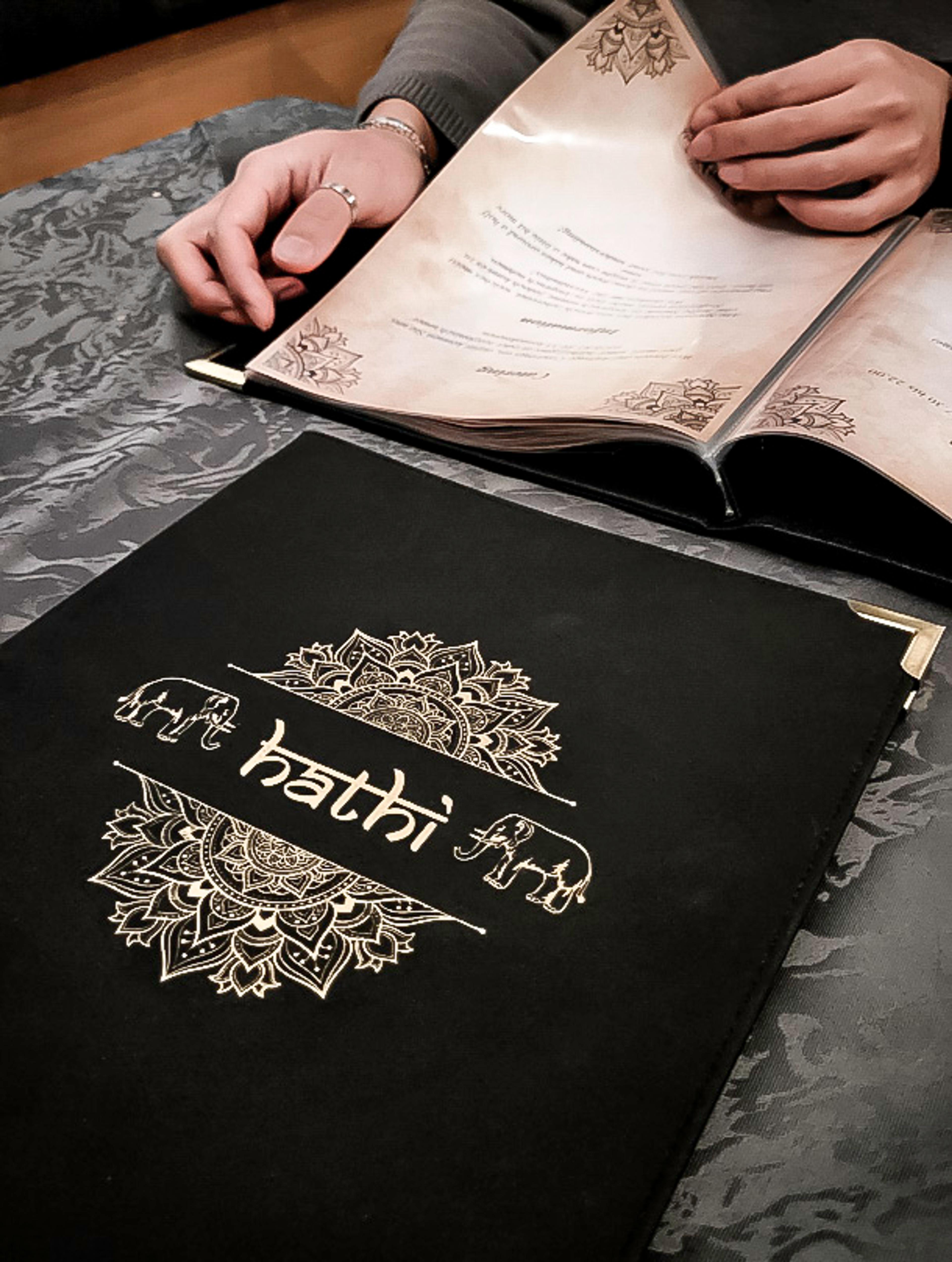 Luxuriöse Speisekarte des Hathi indischen Restaurants mit aufwendiger goldener Prägung auf schwarzem Stoffeinband. Eine Person blättert durch das Menü mit kunstvollen Mandala-Elementen. Edles Branding für exklusive Gastronomie, umgesetzt von Unerwartet Branding und Design OG aus Graz, Österreich.