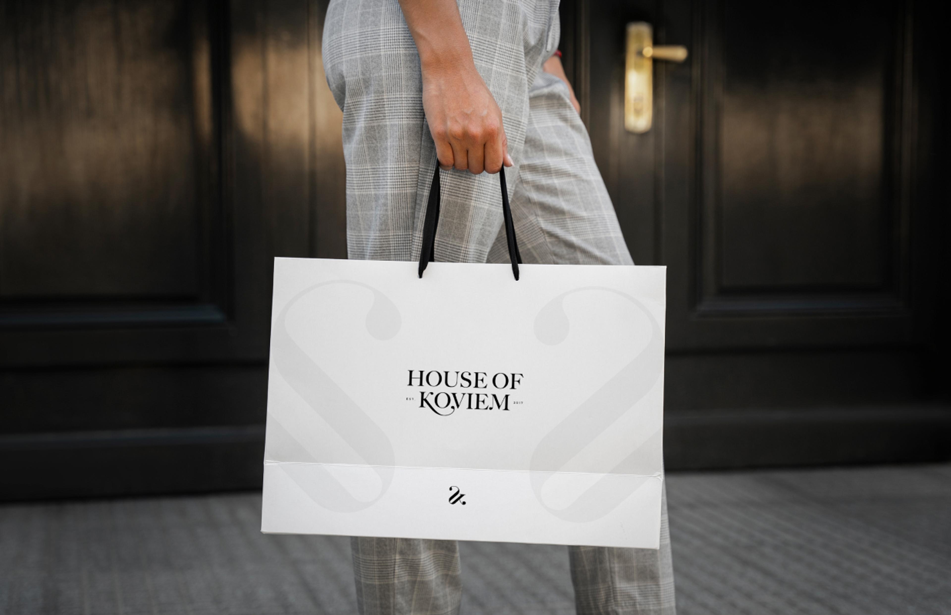 Eine stilvolle weiße Papiertasche mit schwarzem Logo und minimalistischem Design, getragen von einer Person in grauer Hose vor dunkler Tür.