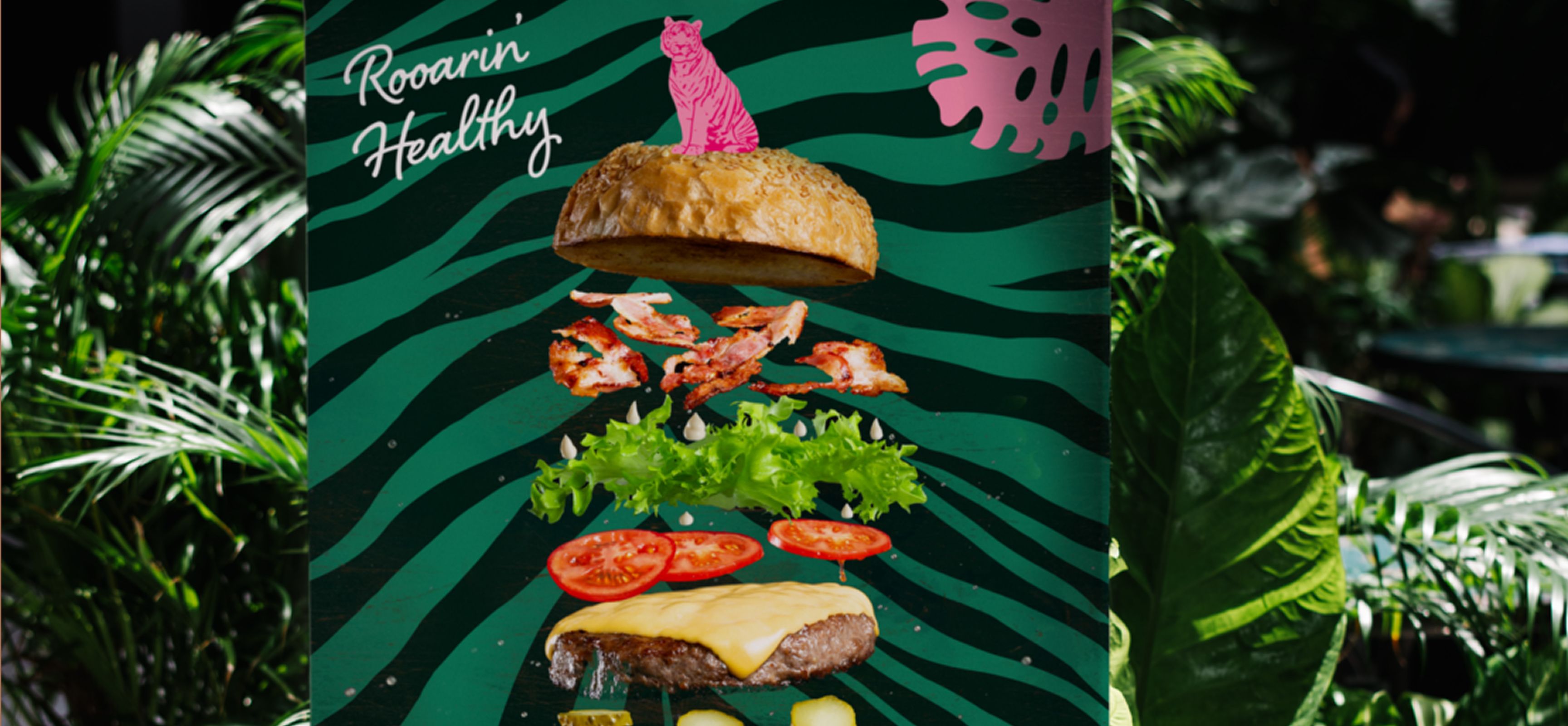 Ein explodierter Burger mit Zutaten in der Luft, auf einem grünen Zebramuster-Hintergrund mit dem Schriftzug „Roarin’ Healthy“.