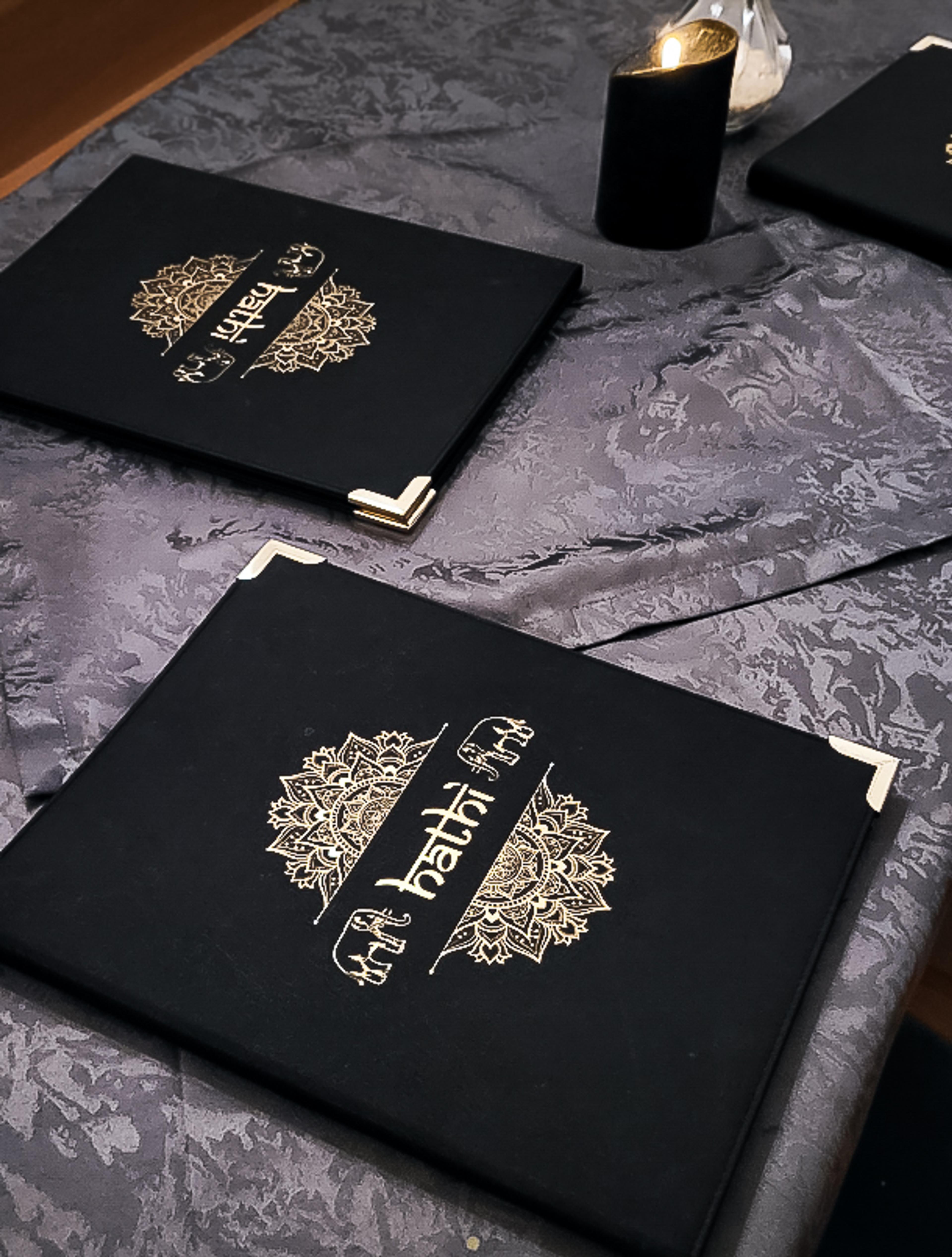 Luxuriöse Speisekarte des Hathi indischen Restaurants mit aufwendiger goldener Prägung auf schwarzem Stoffeinband. Eine Person blättert durch das Menü mit kunstvollen Mandala-Elementen. Edles Branding für exklusive Gastronomie, umgesetzt von Unerwartet Branding und Design OG aus Graz, Österreich.