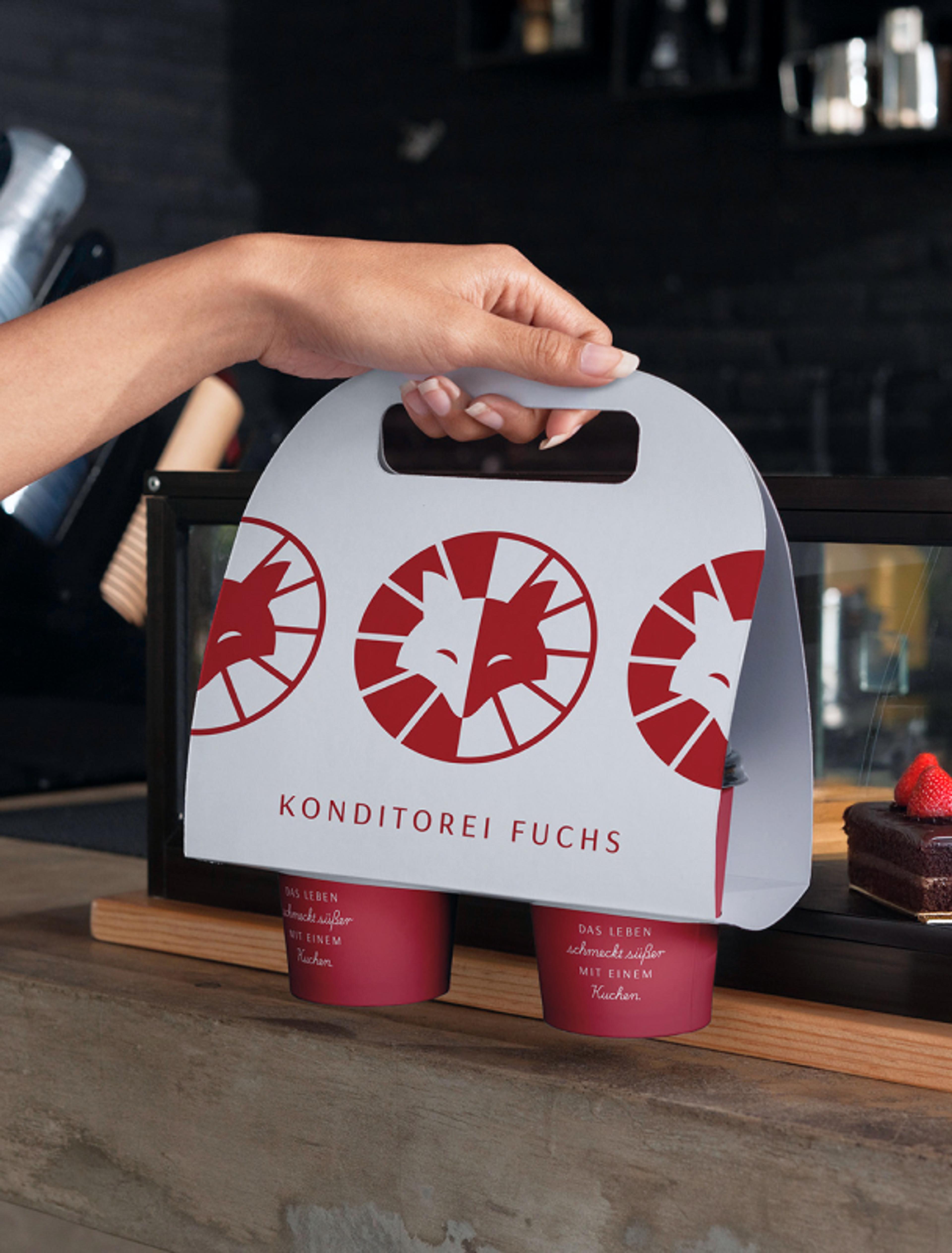 Eine stilvolle, weiße Getränketrägerbox mit dem rot-weißen „Fuchs“-Logo und dem Slogan „Das Leben schmeckt süßer mit einem Kuchen“. Die Box wird in einer Konditorei über die Theke gereicht.
