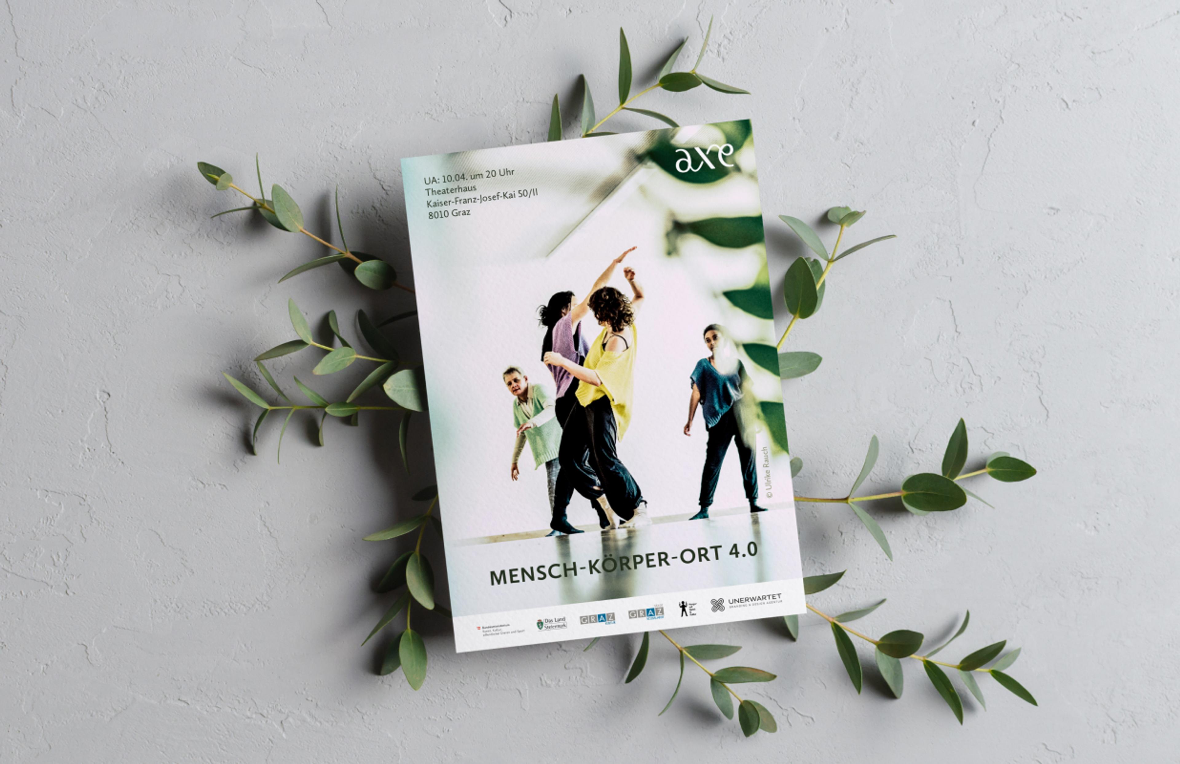 Flyer "Mensch-Körper-Ort 4.0" auf Betonhintergrund mit Pflanzen – Ein Flyer mit tanzenden Personen, arrangiert auf einer Betonfläche mit grünen Zweigen.