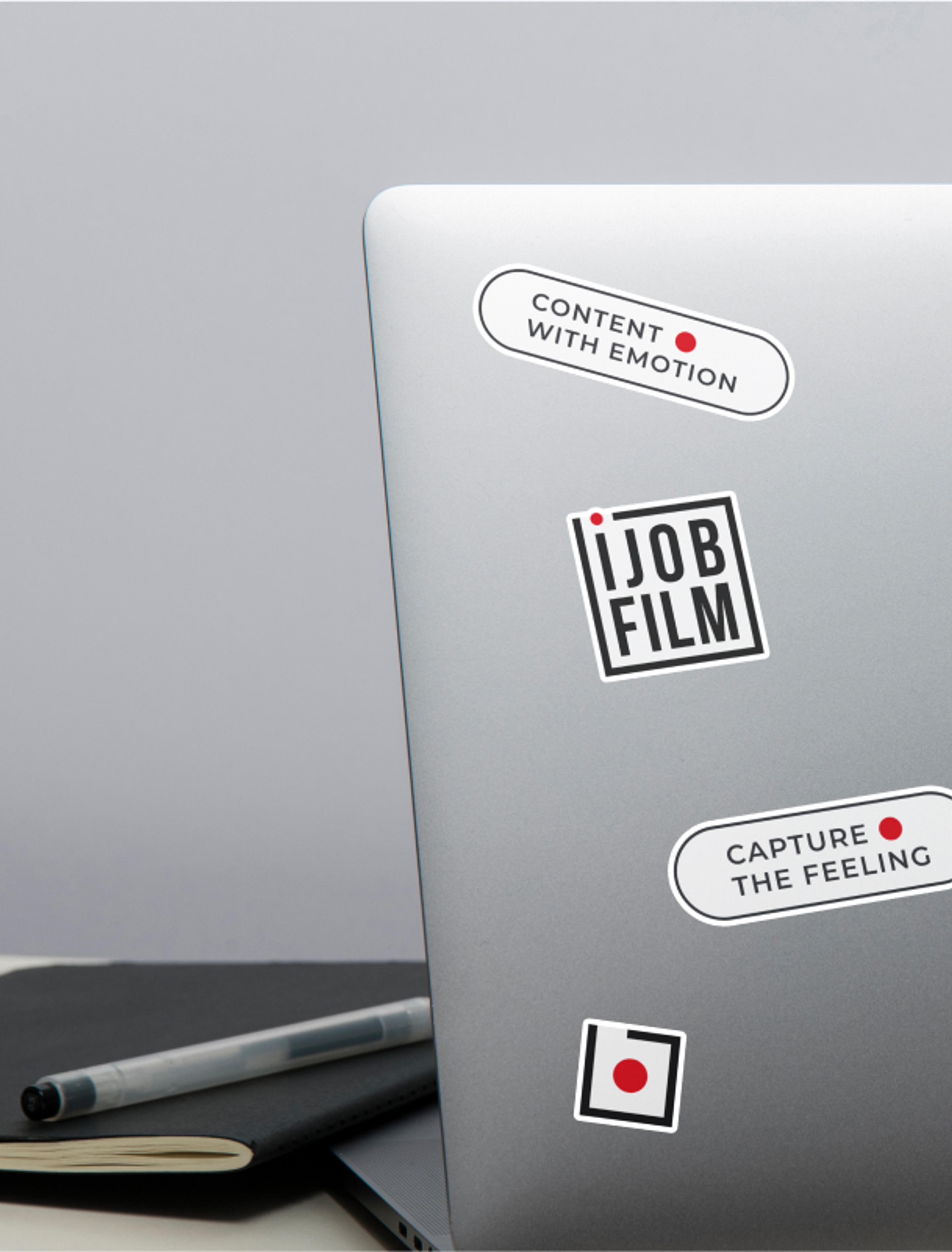 Ijob-Branding auf Laptop-Stickern – Ein Laptop mit mehreren Stickern, darunter das Ijob Film-Logo, der Slogan „Capture the Feeling“ und „Content with Emotion“, alle im typischen Schwarz-Weiß-Rot-Design.