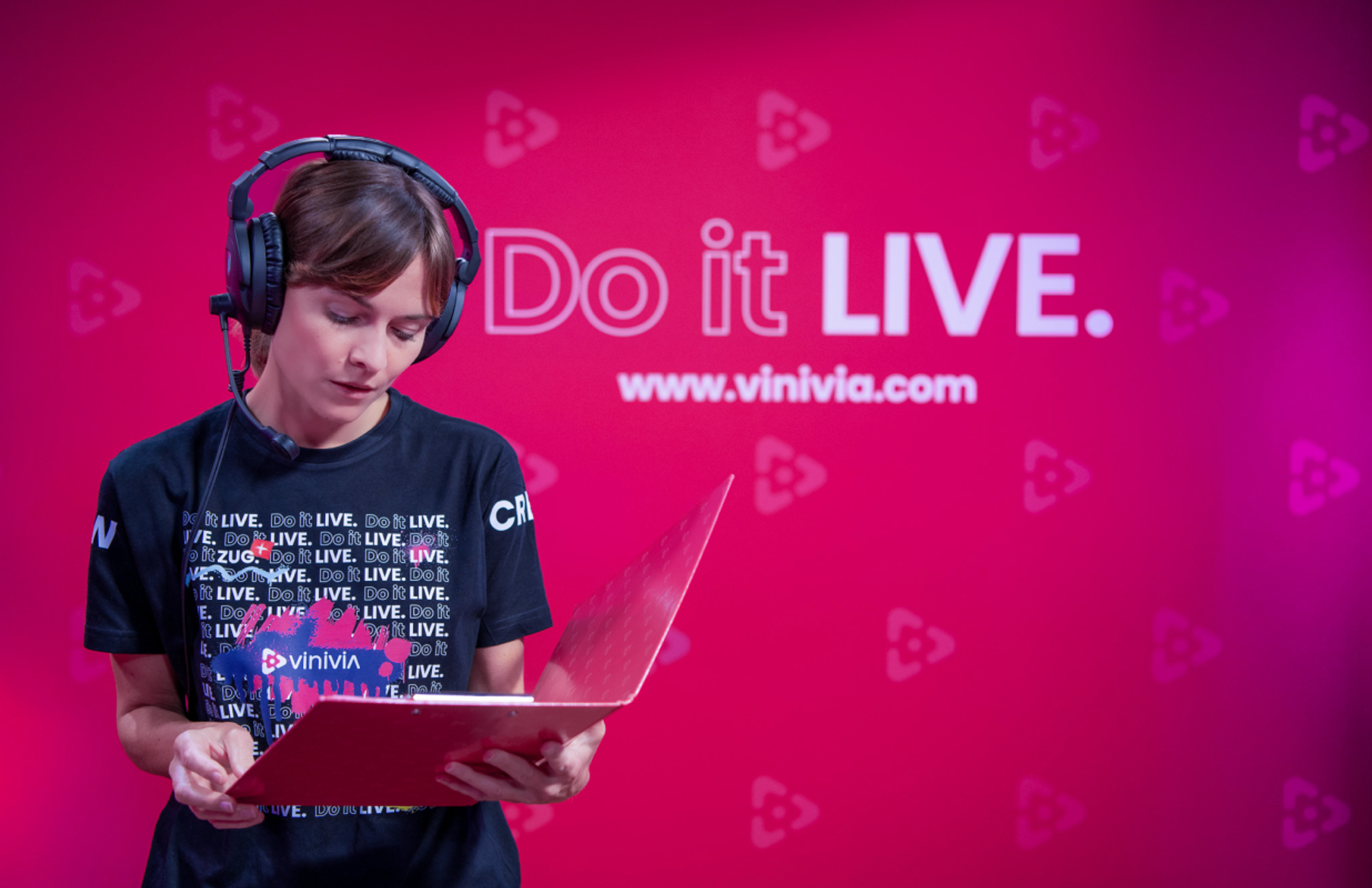 Foto: Vinivia Vinivia-Teammitglied mit Headset und Dokumenten vor einer pinken Markenwand mit „Do it LIVE“-Slogan, symbolisch für professionelle Live-Streaming-Events – Unerwartet Design